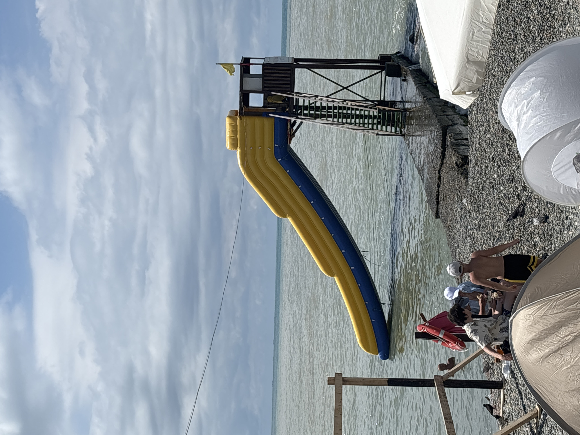 The image depicts a beach scene with a prominent inflatable water slide extending from a wooden platform into the sea. The slide is yellow and blue. A small lifeguard-style tower with stairs is attached to the platform. Several people are visible on the beach and near the water. In the foreground, a beige tent is set up, and a white tent-like structure is partially visible to the right. The beach is covered in pebbles. The sea is choppy under a cloudy sky. A red lifebuoy is attached to a wooden post near the water's edge. The scene is consistent with a recreational area found along the Black Sea coast in Sochi, Russia.