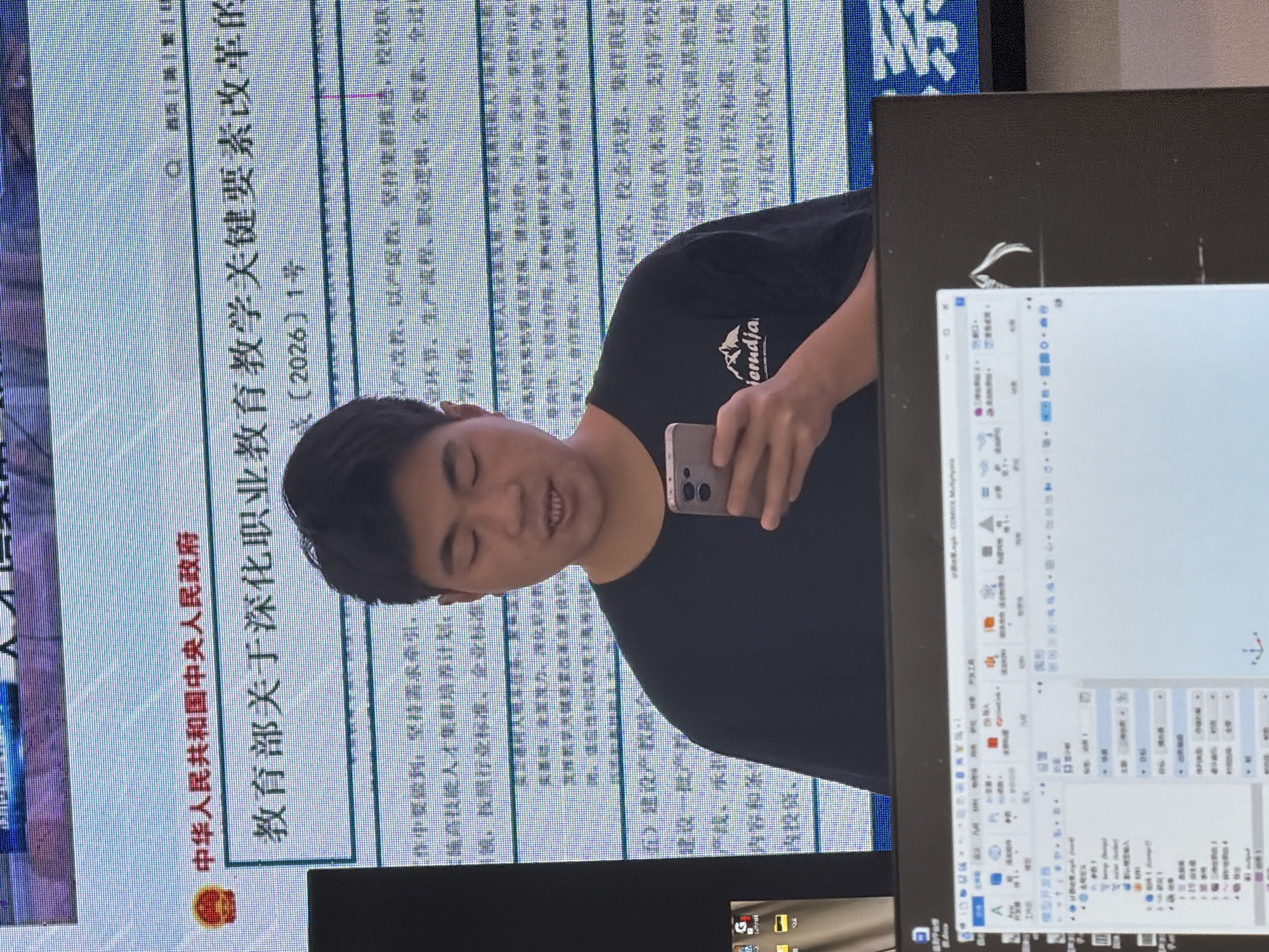 A young man wearing a black t-shirt is standing in front of a large screen displaying text. He is holding a smartphone in his right hand and appears to be looking at it. The screen behind him shows a document with Chinese characters. The man is positioned in the center of the frame. The lower part of the image shows a computer monitor with a software interface open. The screen is displaying a detailed interface with various menus and icons, suggesting a complex application is in use. The overall environment appears to be an indoor setting, likely a classroom or an office, given the presence of the large display screen and the computer monitor. There is no discernible indication of the city of Jiangjin, China in the image itself.