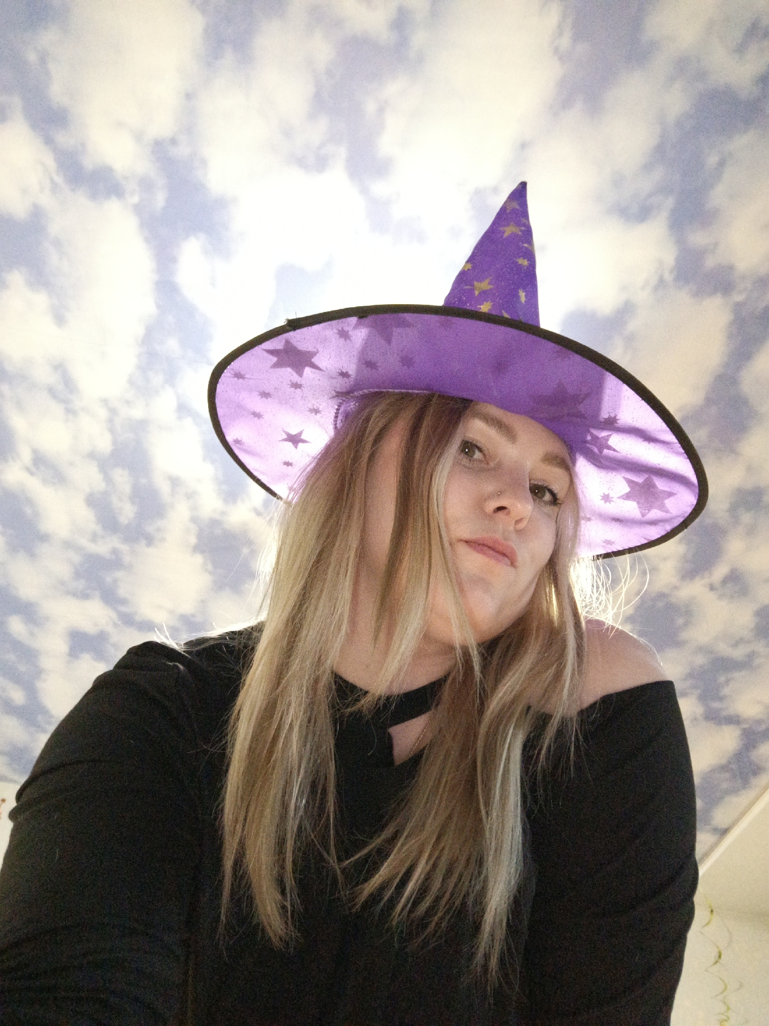 A low-angle shot of a female wearing a purple witch hat with black trim and gold star decals, along with a black off-the-shoulder top. Her hair is blonde, and her face is slightly angled towards the camera. The background is a patterned ceiling, resembling a cloudy sky. The image does not provide enough information to confirm it was taken in Марковский, Russia.