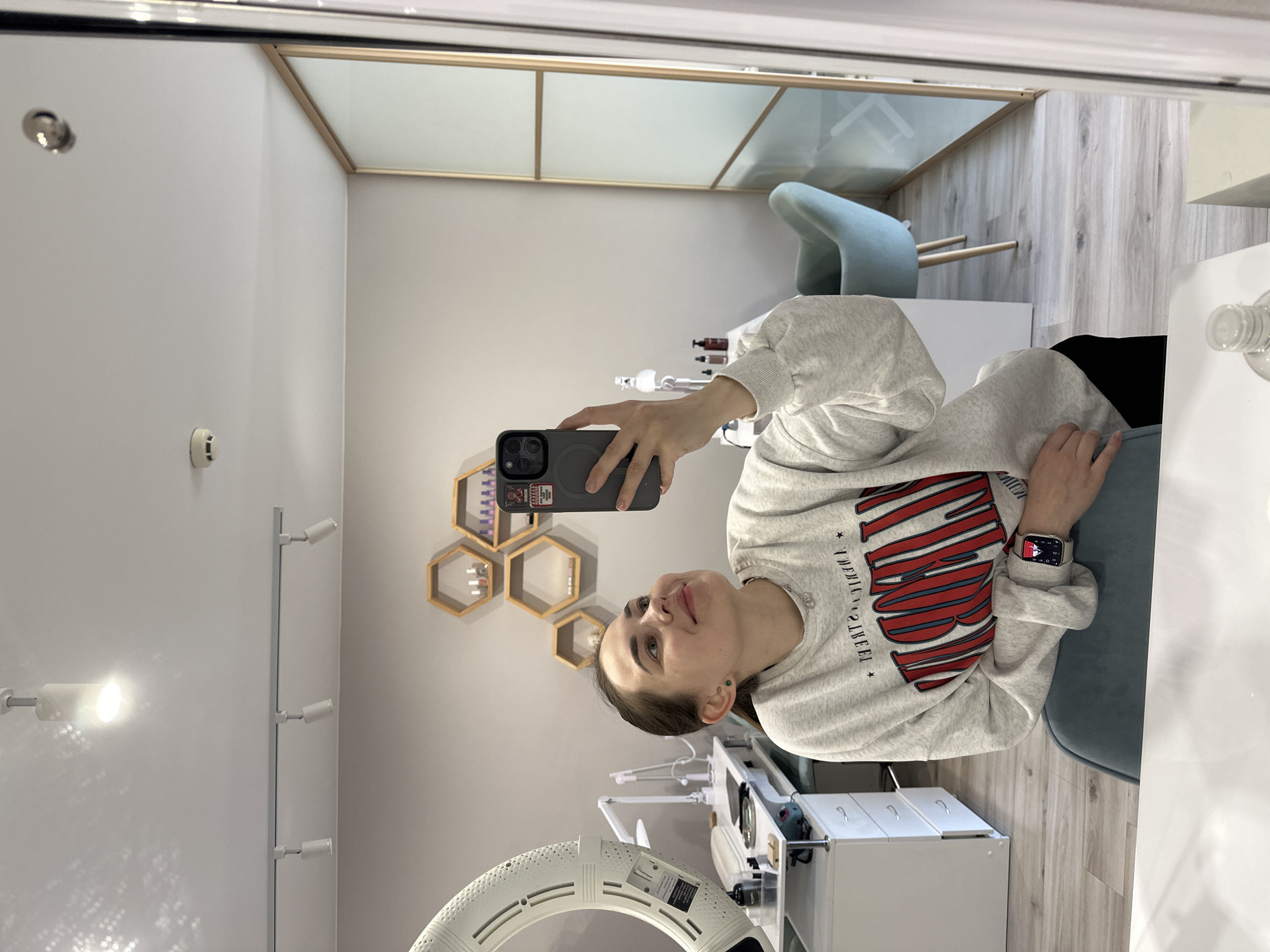 Here is a detailed description of the image:
**Scene:**
The image appears to be a reflection in a mirror, possibly taken in a beauty salon or similar establishment. The setting seems to be in a relatively modern and well-lit room.
**People and Subjects:**
A woman is visible taking a selfie in the mirror. She is wearing a light-colored sweatshirt with red and black lettering/graphics. She has a smart watch on her left wrist.
**Objects:**
* **Room/Interior:** The background includes elements like a partition with frosted glass panels and wooden frames, light-colored flooring (possibly wood or laminate), a light blue chair, and white furniture/cabinets. There are also decorative hexagon shelves on the wall.
* **Equipment:** A ring light is visible in the lower part of the frame. There's also equipment used for beauty treatments/procedures.
* **Electronics:** The woman is holding a smartphone to take the selfie.
* **Wall Fixtures:** A track lighting system runs along the wall.
* **Ceiling:** The presence of a ceiling-mounted smoke detector.
**Actions and Interactions:**
The primary action is the woman taking a selfie. Her posture suggests she is relaxed and possibly happy.
**Environment:**
The room is clean, orderly, and professionally designed, which supports the idea that it is a beauty salon or similar commercial space.
**Location:**
The available information suggests the location is in the city of Perm, Russia. However, this information is not discernable from the image itself, but based on the prompt.