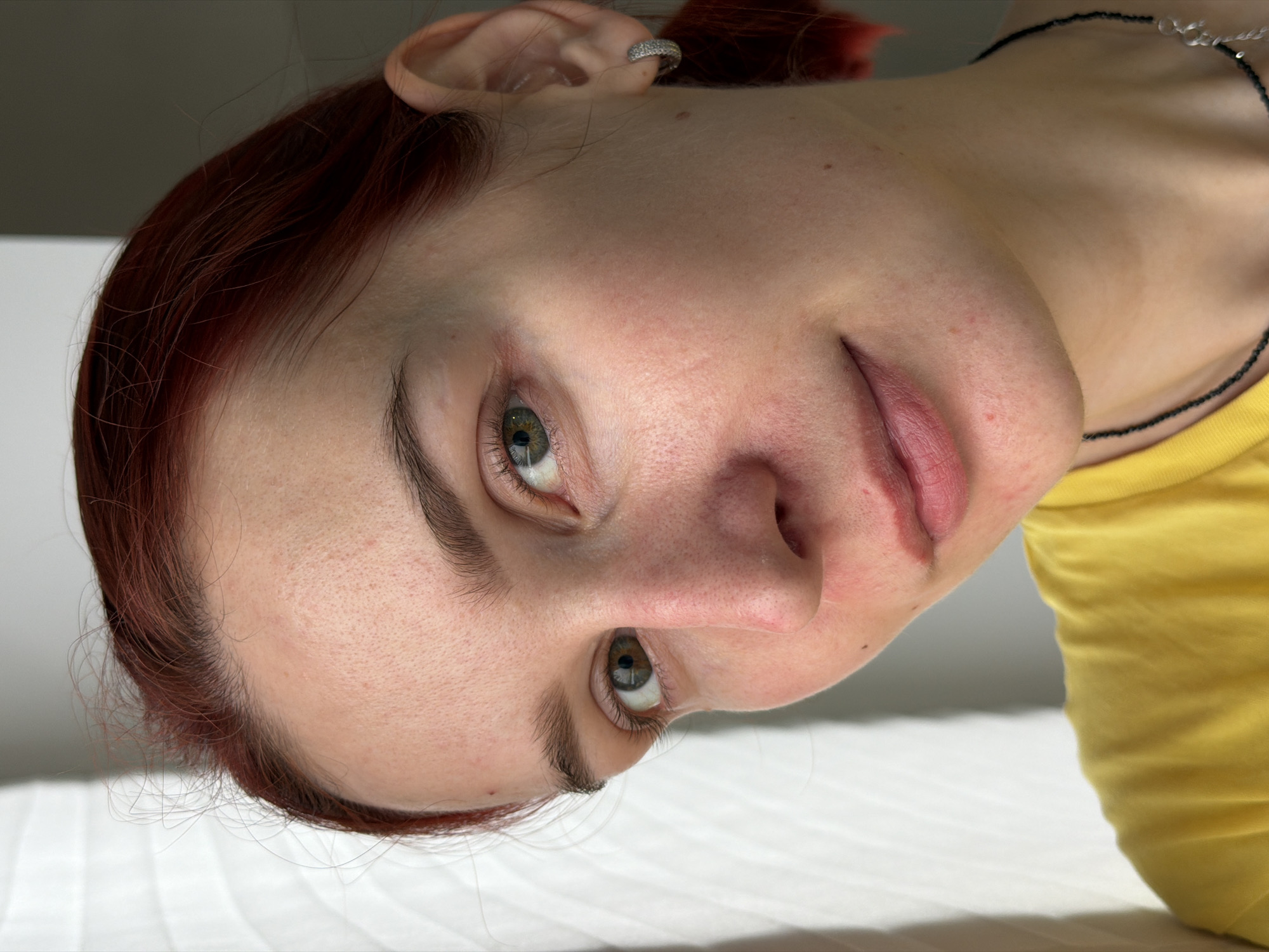 A close-up, low-angle, tilted shot features a fair-skinned woman with red hair. Her eyes are green, and she has a silver hoop earring in her left ear. The woman is wearing a yellow shirt and a black necklace with silver accents. The background is a mix of white and gray, suggestive of a bed and wall. No specific details indicate the location of Chaikovsky, Russia.