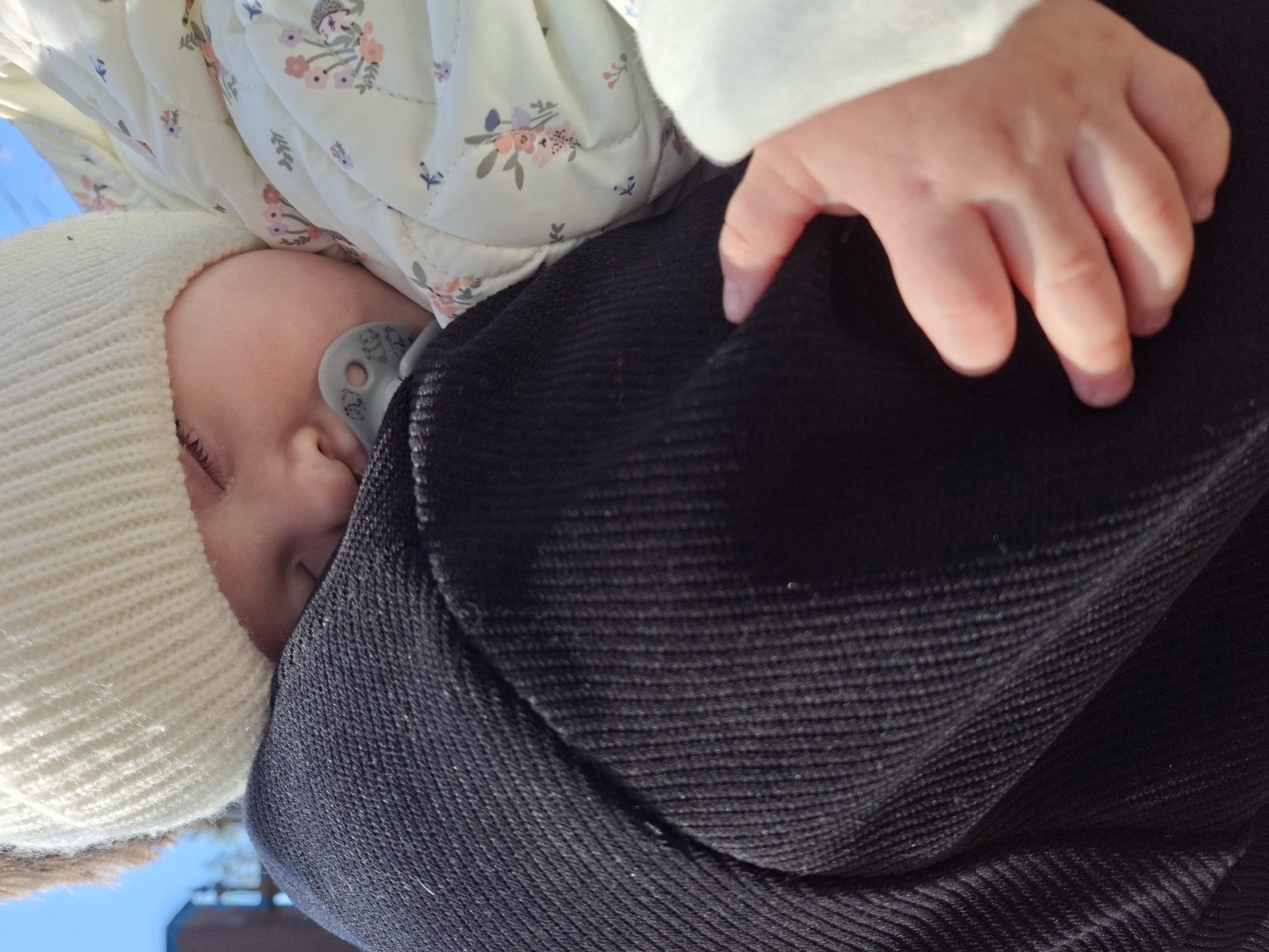 A close-up shot shows an infant being held, seemingly outdoors. The infant is wearing a white knit cap and a floral-patterned jacket, and has a pacifier in their mouth. The child is being held against a dark-colored fabric, possibly a coat or blanket, with a small hand resting on top. The image seems to be taken from a low angle, with a glimpse of the sky and what may be a building in the background. Without further information, it's not possible to confirm the location as Chaikovsky, Russia or any further details about the environment.