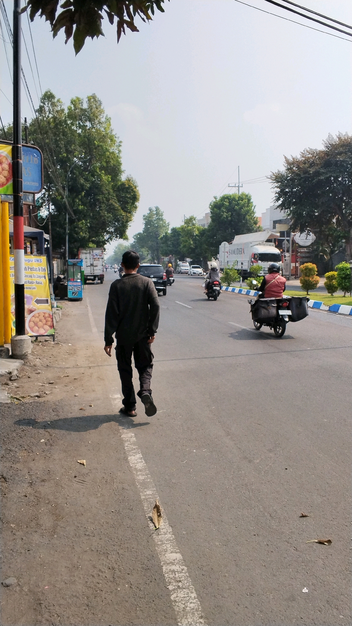 A man walks on the side of a road in Banjaragung, Indonesia, away from the camera. He wears a dark long-sleeved shirt and dark pants, with his left arm slightly extended. The road has a white dashed line on the right side of the lane he is walking parallel to. On the left side of the road is a sidewalk with a pole and several signs, one of which displays text in Indonesian and an image of food. Further down the road, other vehicles are visible, including cars, a white truck with "SAMUDERA" written on it, and several motorcycles. The scene appears to be in a populated area with trees lining the opposite side of the road and buildings in the background. The sky is bright and hazy, suggesting a sunny day.