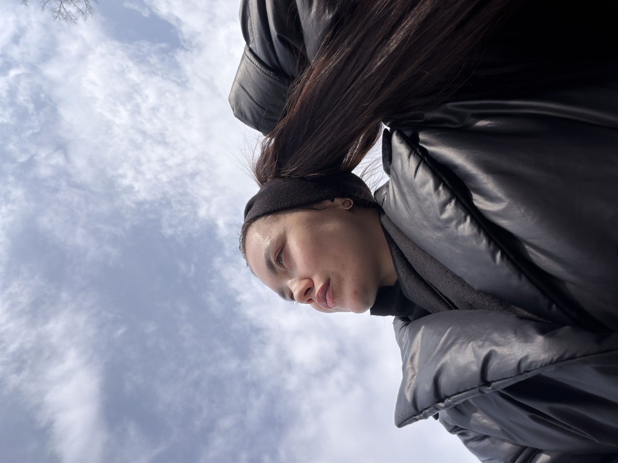 A low-angle shot depicts a person with brown hair, wearing a black puffer jacket and a black headband, against a cloudy sky. The person is positioned in the upper right of the frame, while the sky occupies the lower left. The photograph was taken in the city of Chaikovsky, Russia.