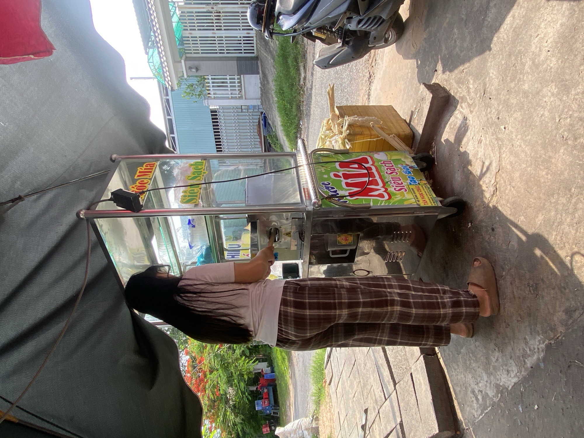 A woman with long dark hair, wearing a light pink t-shirt and brown plaid pajama pants, stands facing a metal cart under a dark green awning. The cart has glass panels and a stainless steel exterior, with a logo that includes the word "MÍA" in red and yellow. Inside the cart, various items are visible, including plastic containers. The woman appears to be operating a device on the cart, holding a stick-like object. In front of the cart, a wooden box contains sugarcane stalks. To the right of the cart, a dark-colored motorcycle is parked, with a black helmet on its handlebars. The ground is made of concrete slabs and cracked concrete. In the background, there is a white fence, a building with blue walls, and some trees with red flowers. The overall setting suggests an outdoor stall in Mũi Né, Vietnam, likely selling sugarcane juice.