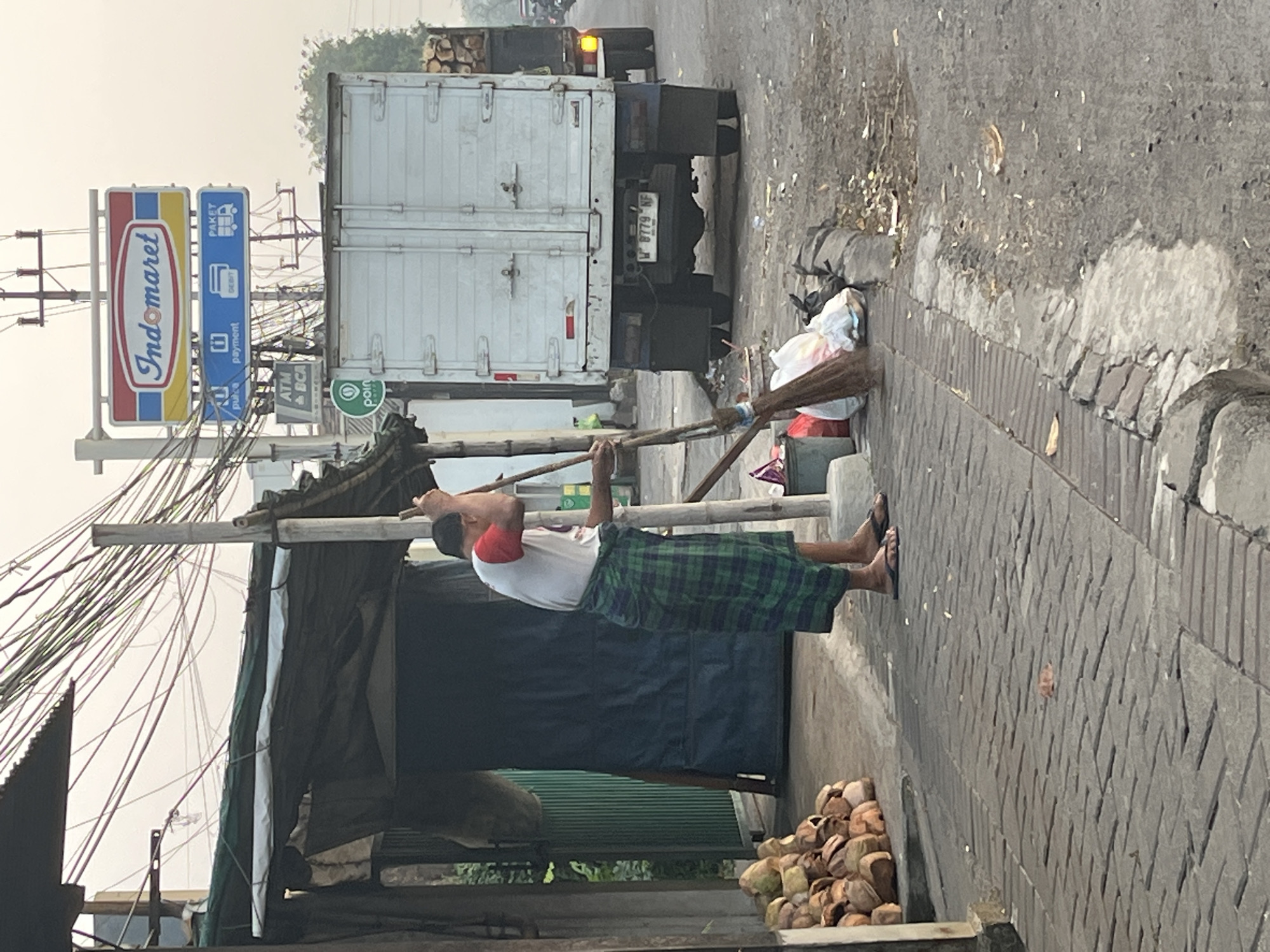 A man is sweeping the sidewalk in front of a small shop in Kludan, Indonesia. He is wearing a red and white shirt and a green and blue plaid sarong. He is holding a broom in his right hand and is sweeping towards the street. In the background, there is a white truck with its rear doors open, and a pile of coconuts is stacked on the left side of the image. There are also utility wires overhead and a sign for "Indomaret" in the background. The sidewalk is made of brick and the street is paved with asphalt. The scene appears to be in a commercial area, with shops and other businesses lining the street. The image is taken from a slightly low angle, looking up towards the man and the truck.