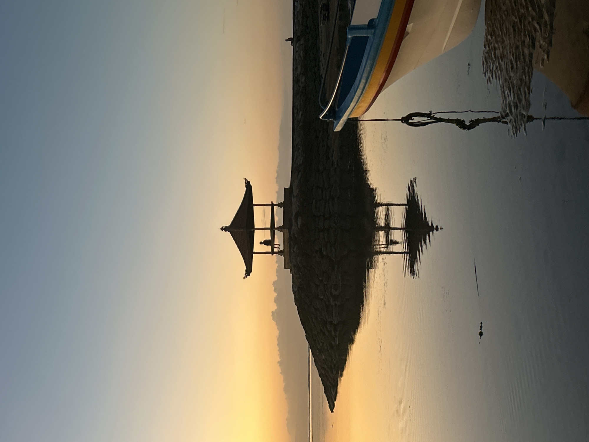 At dawn in Denpasar, Indonesia, a traditional Balinese gazebo stands on a rocky outcrop beside calm water. A lone figure is silhouetted inside the gazebo, seated on a bench. The sky displays a gradient of soft yellow and pale blue, with a line of clouds near the horizon. The gazebo and its reflection are clearly visible in the still water, which mirrors the warm tones of the sky. In the foreground, the bow of a white boat with blue and yellow trim rests near the shore, its rope tied and dangling in the water. The reflection of the rope is also visible. The overall scene evokes a sense of tranquility and quietude.