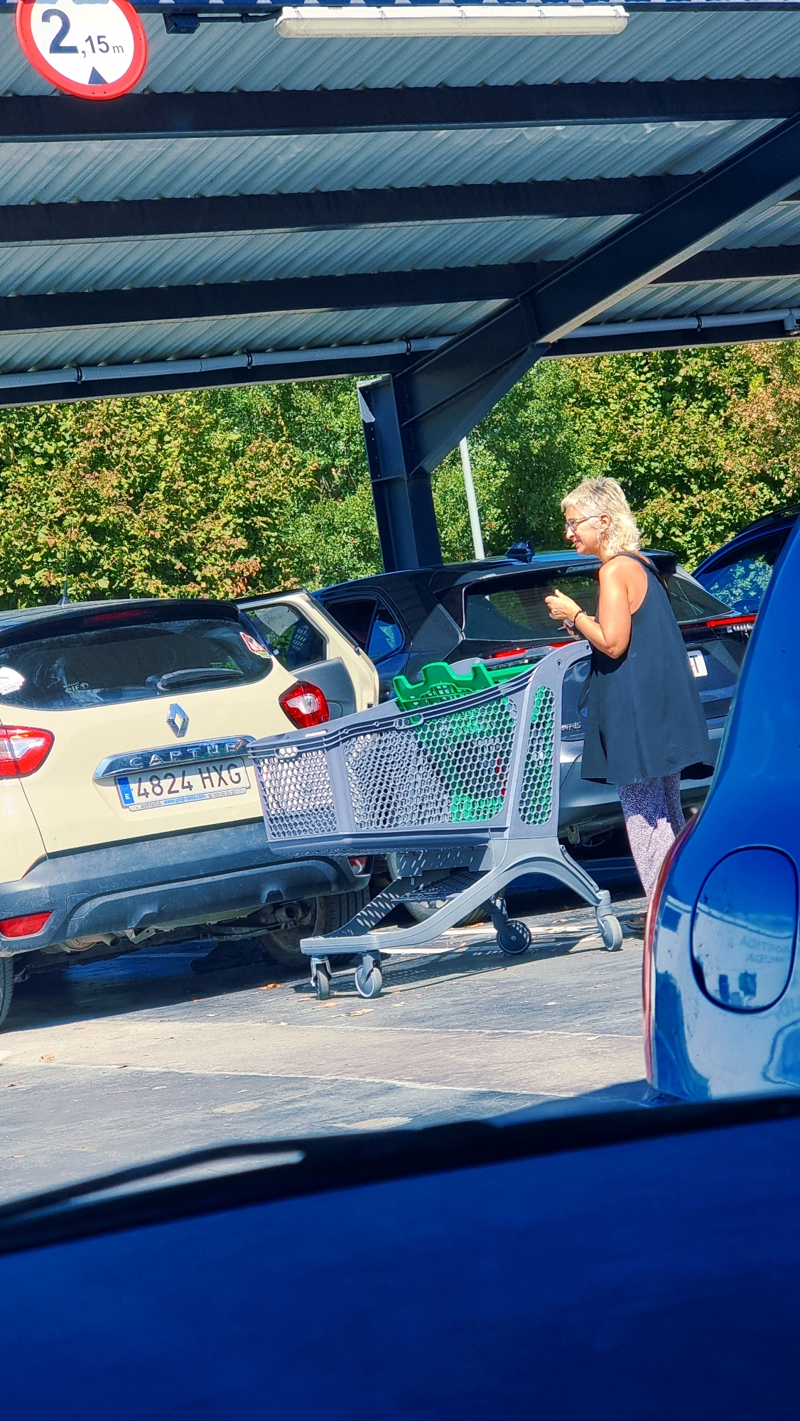 A blonde woman in a black sleeveless dress and patterned leggings stands in a parking lot, pushing a shopping cart. The cart contains two green plastic baskets. Behind her, a light-colored SUV with the license plate "4824 HXG" is parked. Further back, a dark-colored car is visible. A sign indicating a height limit of 2.15 meters is affixed to the covered structure overhead. The environment appears to be a sunny day with trees visible in the background, likely a parking lot in Olot, Spain.