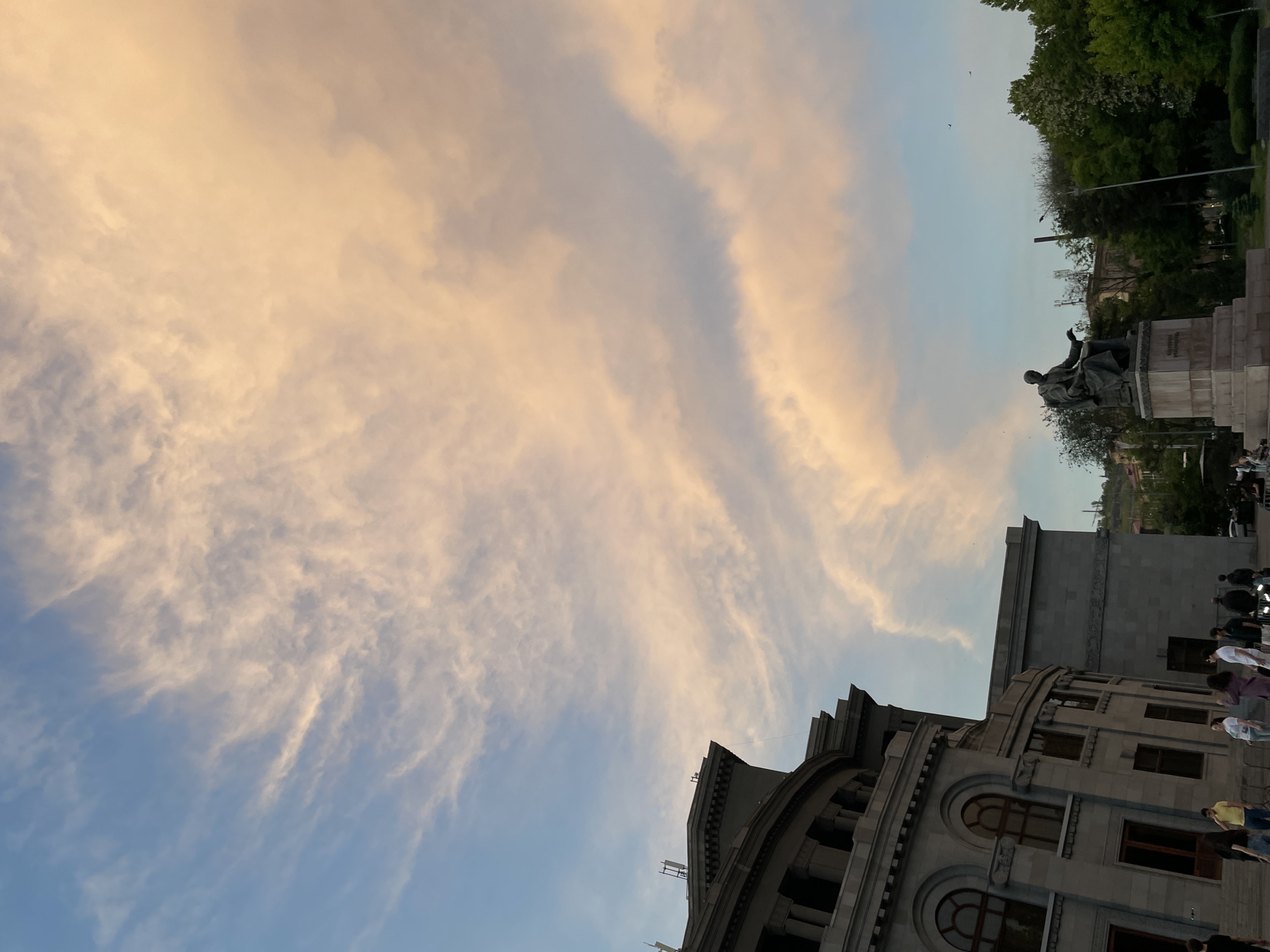 The image captures a view in Yerevan, Armenia, showcasing the sky, a building, and a monument. In the upper-right, a statue depicting a seated male figure is placed atop a stone pedestal. Adjacent to it is greenery, indicating a park-like setting. Below the statue, a grey, multi-story stone building featuring arched windows and intricate architectural details is visible. A group of people can be seen near the building's base on the right. Above the building and monument, the sky is filled with elongated, striated clouds tinged with hues of orange and yellow, transitioning into a blue tone near the bottom of the frame.