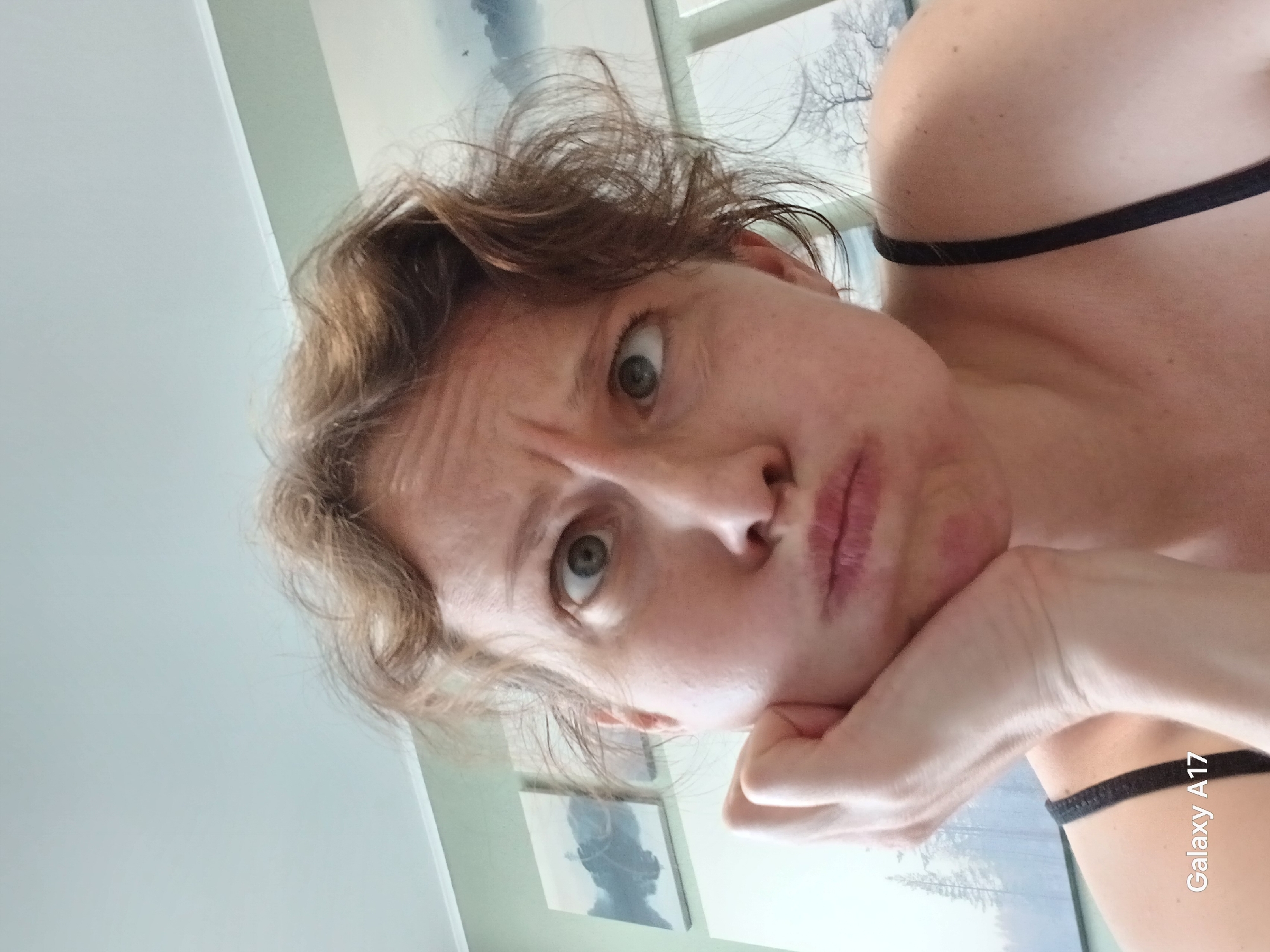 A woman is positioned in a selfie pose with her hand propping up her face. She has fair skin, light brown hair, and green eyes. Her expression appears pensive or slightly questioning. She is wearing a black strap top. There is a painting on the wall behind her and a window. "Galaxy A17" is visible in the bottom right corner of the image. The location in Yekaterinburg, Russia is not determinable from the provided image.