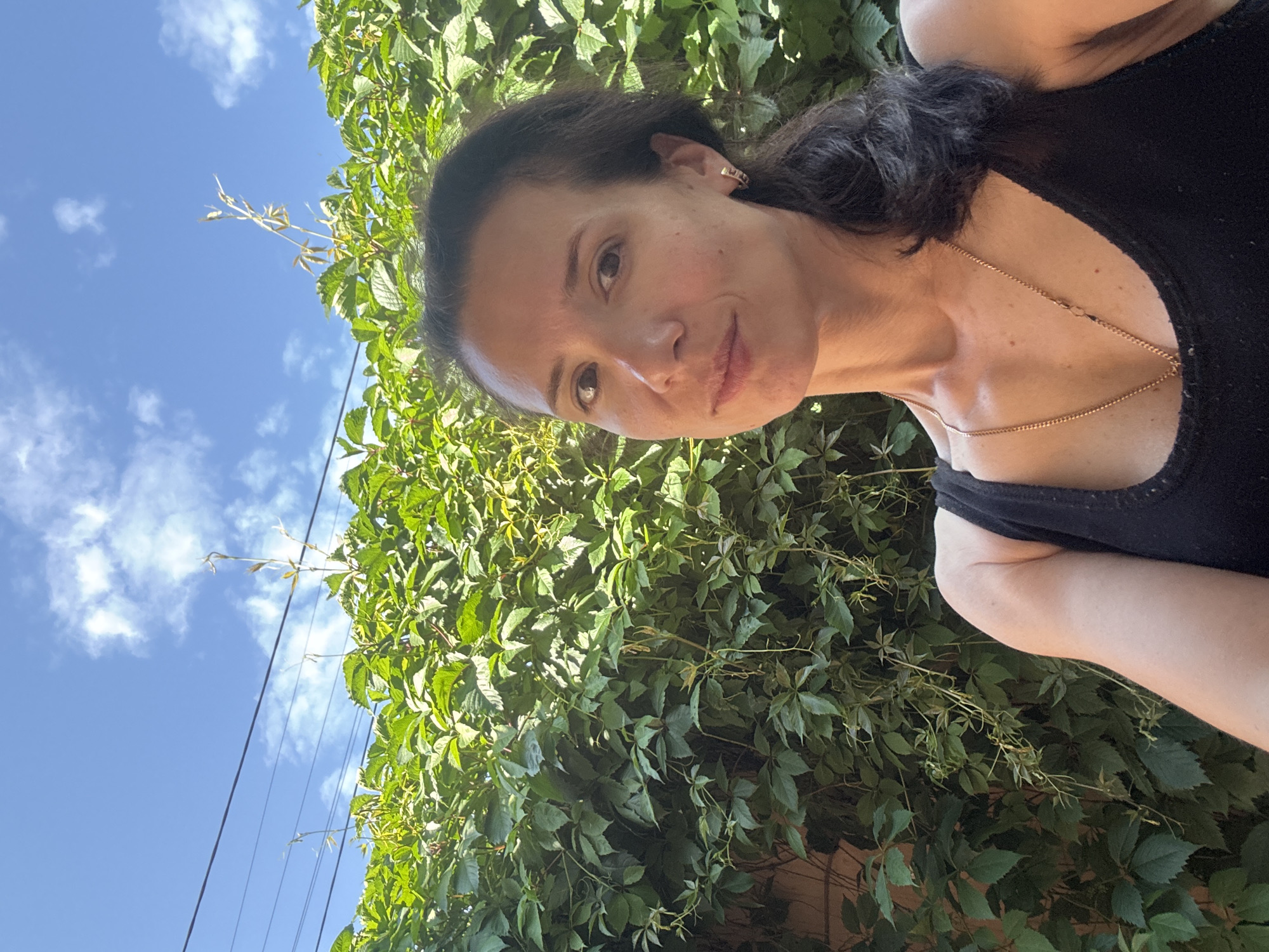 A woman with dark hair pulled into a ponytail stands outdoors, facing the camera. She is wearing a black tank top and a gold necklace. Behind her is a dense green vine-covered wall, and above that, a blue sky with scattered clouds and power lines. The lighting suggests it is daytime.