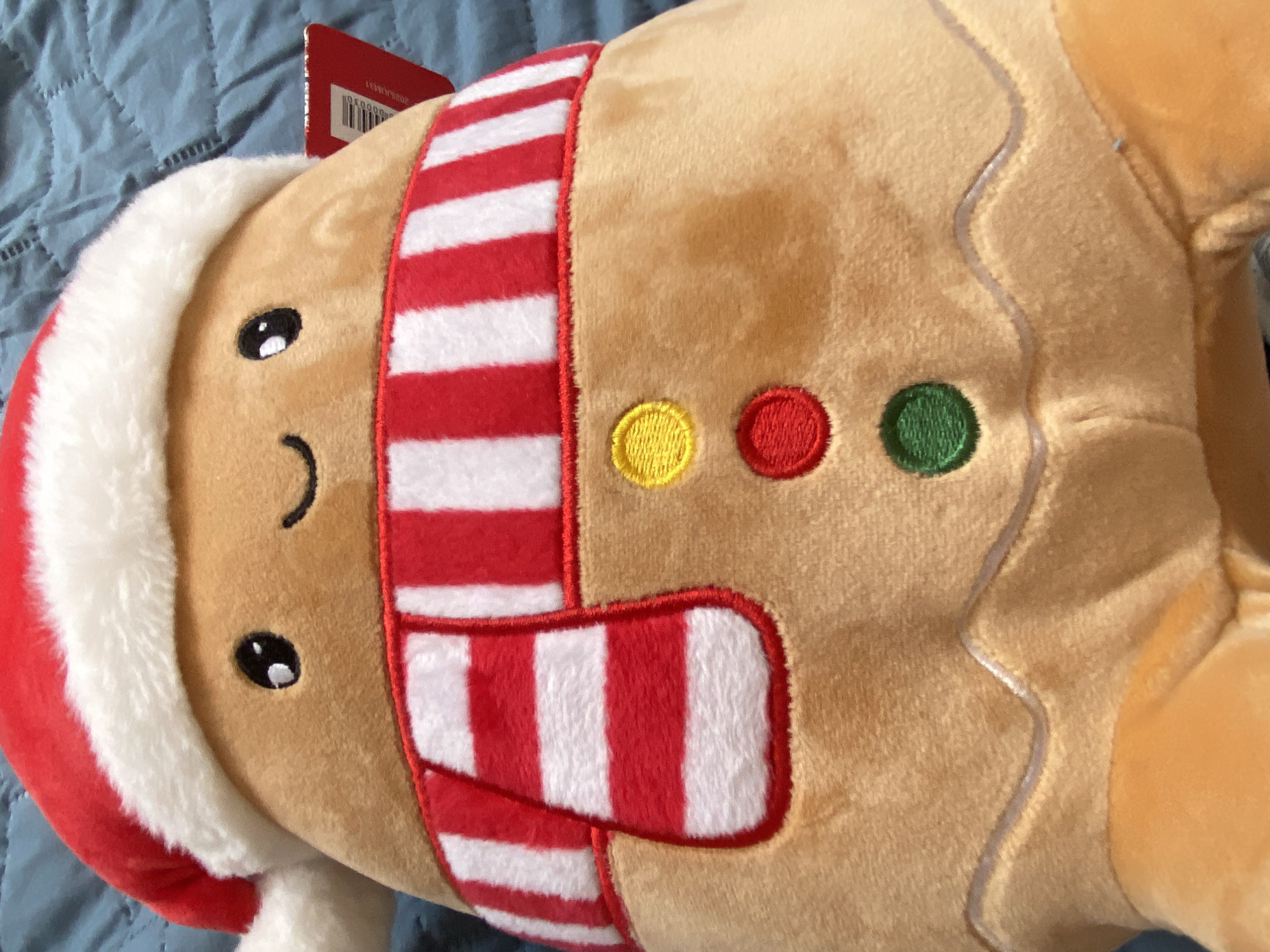 The image shows a close-up of a gingerbread man plush toy. The toy has a tan body with a white fur trim on the top of its head and a red and white striped scarf. It has stitched eyes and a stitched smile. There are three button-like decorations on its body in yellow, red, and green. The background is a light blue fabric. There is no information to associate this image with the city of Konstantinovo, Bulgaria.