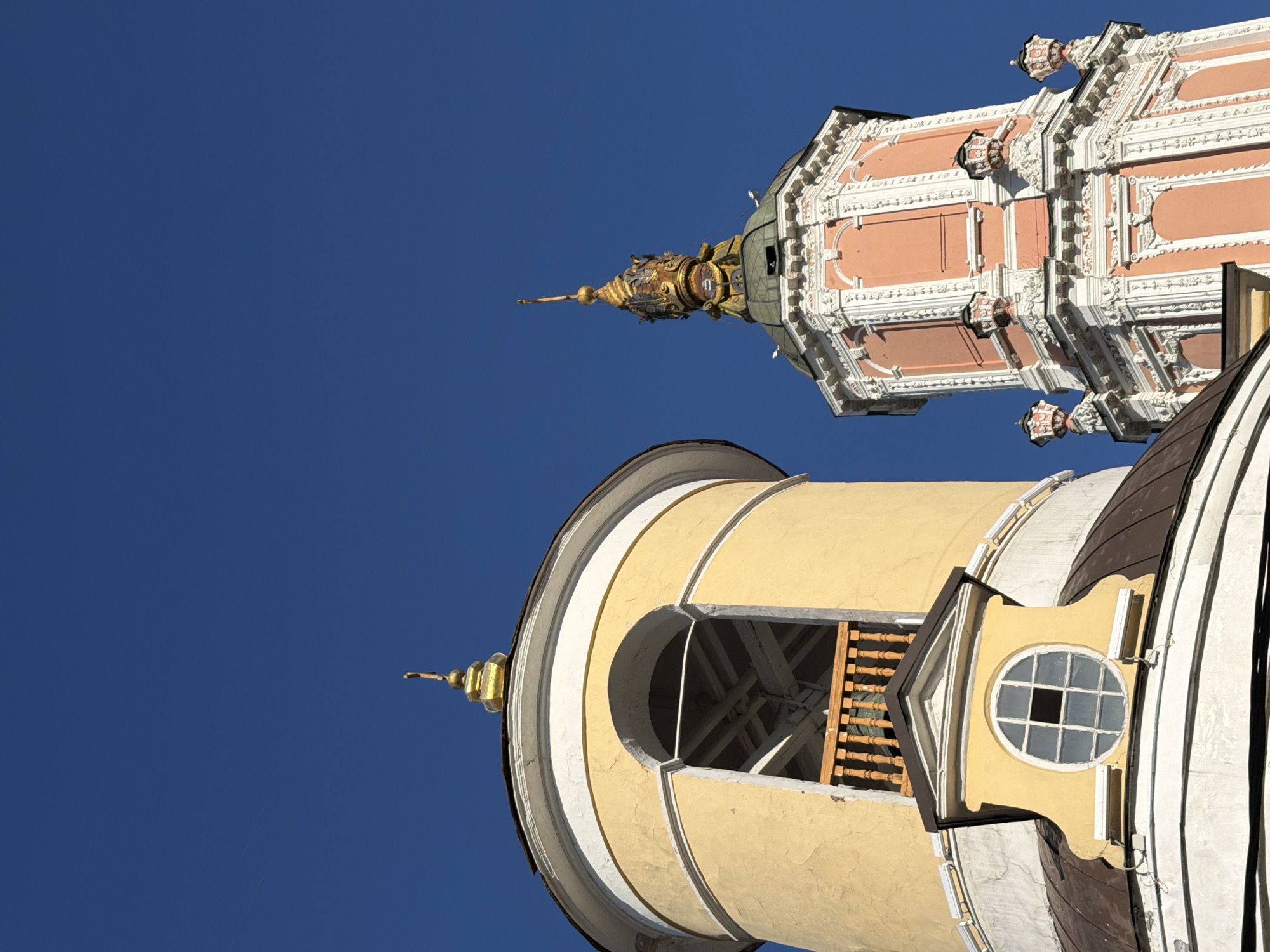 The image shows two towers of the Novo-Devichy Convent in Moscow, Russia, angled upwards against a clear blue sky. The lower tower is cylindrical and colored in shades of cream, white, and brown, featuring a round window with a grid pattern. An open arched section reveals wooden structures. The upper tower is a more elaborate, multi-tiered structure, painted in shades of salmon pink and white, adorned with decorative architectural elements. Both towers are topped with golden spires.