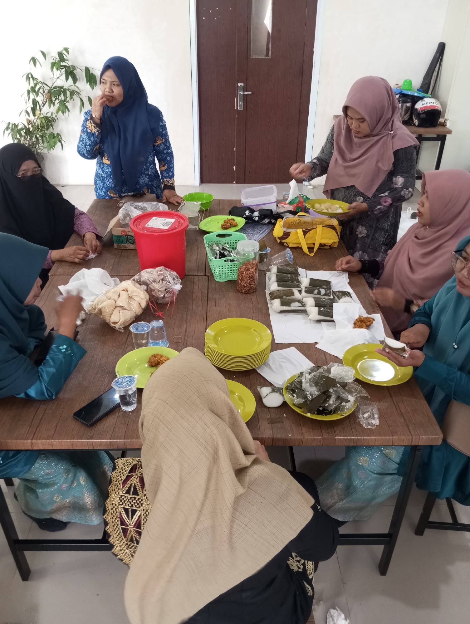 The image depicts a group of five women gathered around a wooden table, likely in a residential setting in Pematang Reba, Indonesia, possibly a community gathering or meal. The women are all wearing hijabs and modest attire, indicating a cultural context common in the region.
In the foreground, a woman in a teal hijab and long blue dress is seated and appears to be holding a piece of food, possibly a snack wrapped in banana leaves, towards her mouth. Beside her, a woman in a beige hijab and dark patterned clothing is also seated, her face obscured by her head covering. Another woman in a dark hijab and black abaya is seated on the left side of the table, partially visible.
Further into the scene, a woman in a dark grey dress and a rose-colored hijab is standing and actively engaged with the food, holding a small plate with a fork. Her facial expression suggests concentration on the food. Another woman in a navy blue hijab and a patterned blue and yellow top is also standing and eating from her hand.
The table is laden with various food items and serving dishes. There are plates stacked, some containing snacks wrapped in leaves, and individual servings of what appear to be fried snacks or pastries. A bright red plastic container is prominent in the center, alongside a green plastic basket filled with utensils. Bags of snacks, possibly crackers or chips, are also present. Several plastic cups, some containing beverages, are scattered across the table.
The environment appears to be an indoor space with white walls and a wooden door in the background. A potted plant with green foliage is situated to the left of the frame. The overall impression is one of a casual social gathering focused on sharing food and conversation.