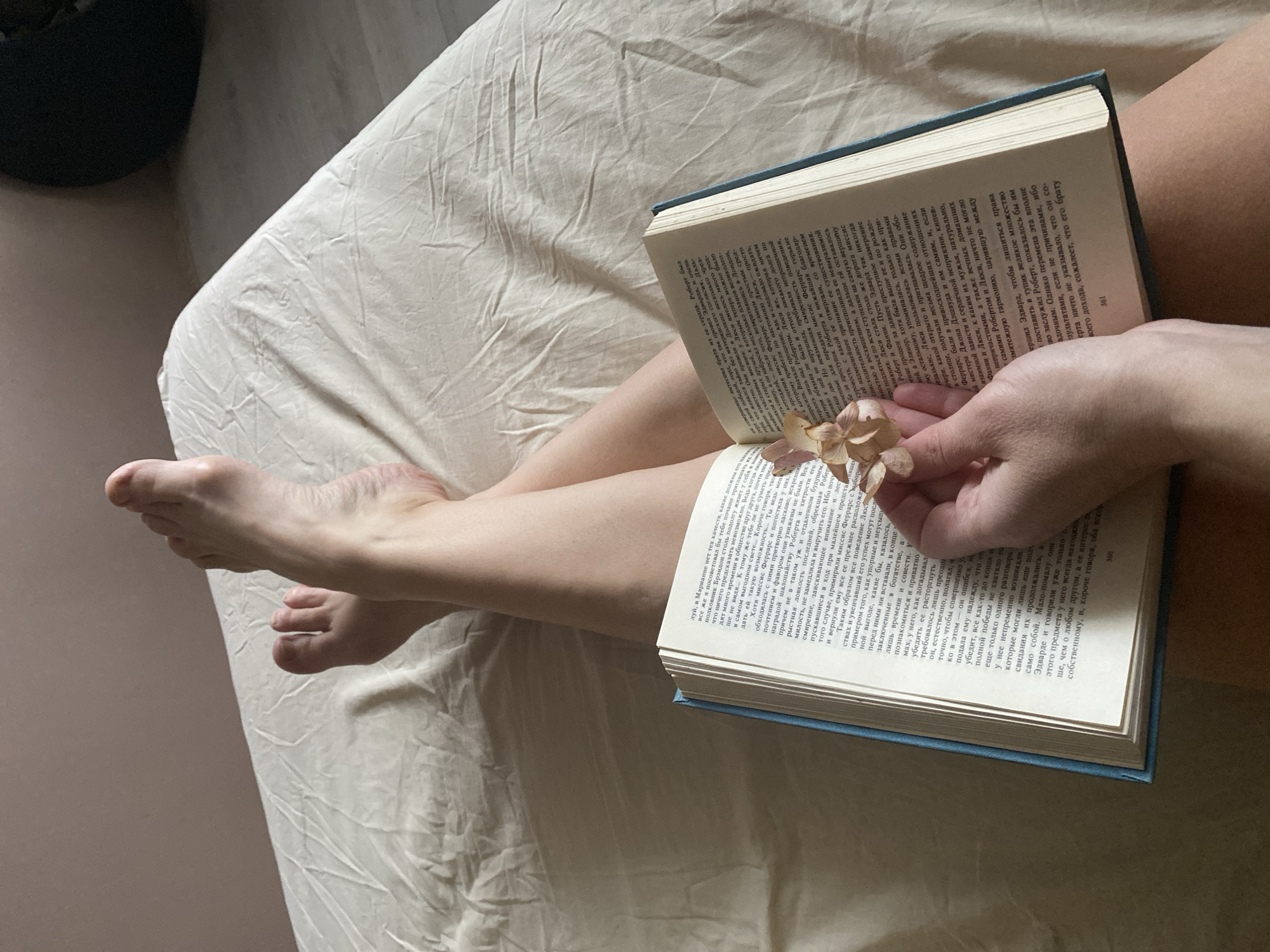 The image shows a close-up of a person lying in bed with an open book. The book is held open with one hand, with a dried flower placed in the middle of the pages. The person's legs and feet are visible on the left side of the frame. The bed has a light-colored, wrinkled sheet. The context of Chaikovsky, Russia is not apparent from the image alone.