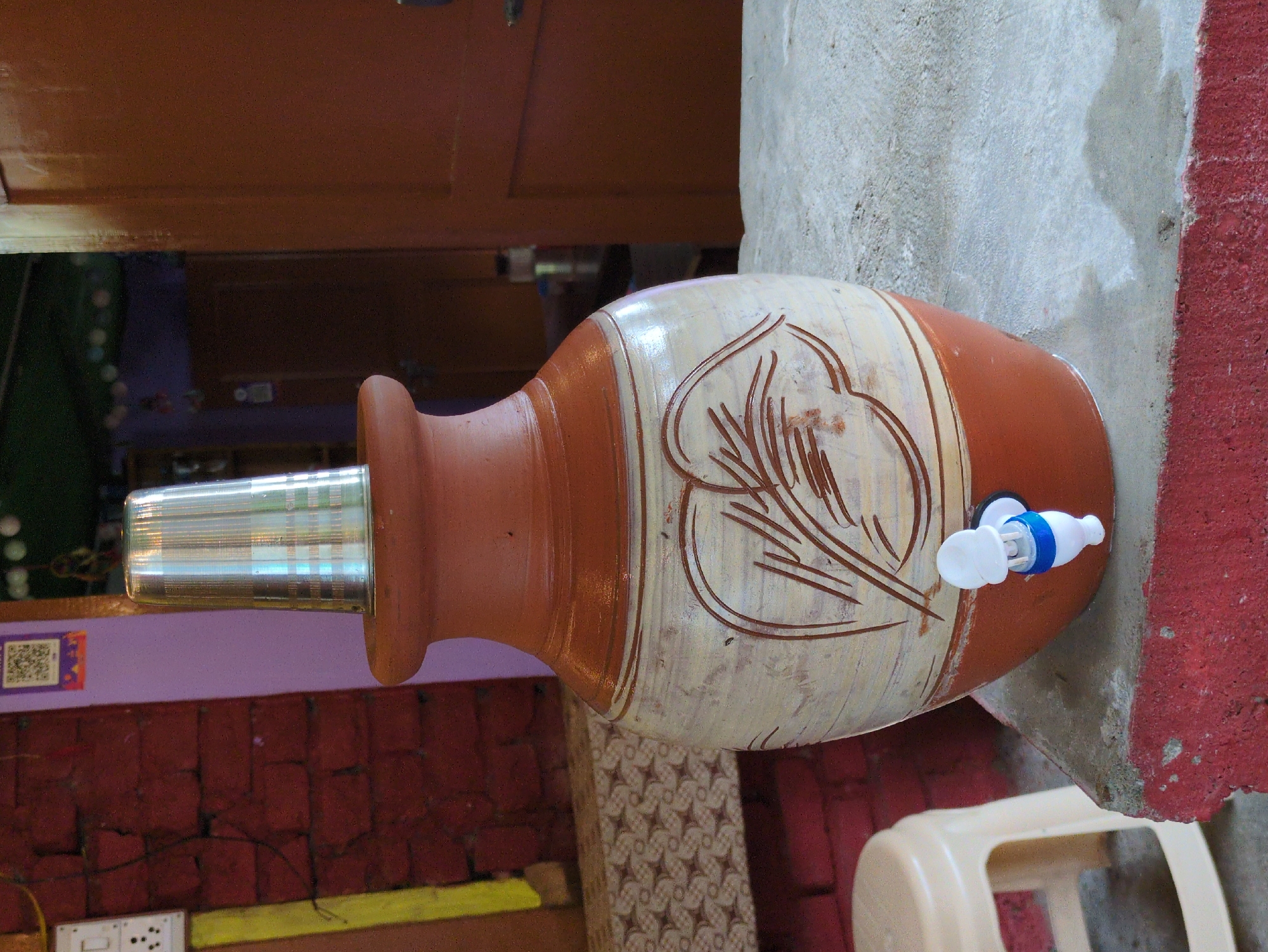 A traditional clay water dispenser with a silver cup on top sits on a concrete ledge. The dispenser is decorated with a cream-colored band featuring a leaf motif in brown. A white and blue spigot is attached to the lower portion. To the left, a small, light-colored plastic stool is visible. In the background, a brick wall with a purple section and a wooden door can be seen. The setting appears to be outdoors, possibly in a courtyard or on a patio.