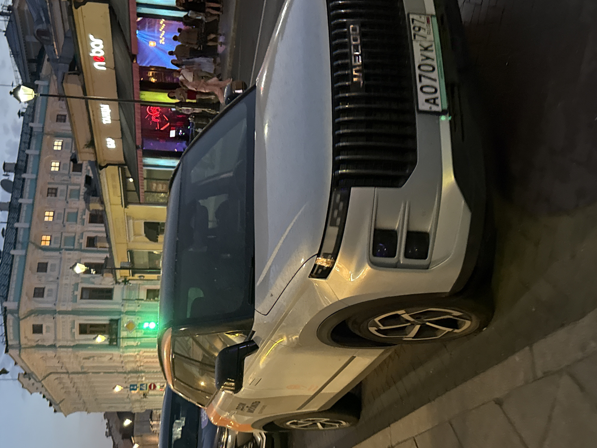 In Moscow, Russia, on a street at dusk, a white SUV with orange accents is parked on the side of the road. The vehicle has a distinctive grille with the "JAECCO" logo. A Russian license plate, "A070YK 797", is visible on the front. Across the street, a building with illuminated windows and ornate architectural details stands prominently. Adjacent to it is a establishment with a neon sign that reads "nōbar" and "GAP TAKHI". Several people are walking on the sidewalk in front of the establishments. The scene is illuminated by streetlights and the glowing signs of the businesses.