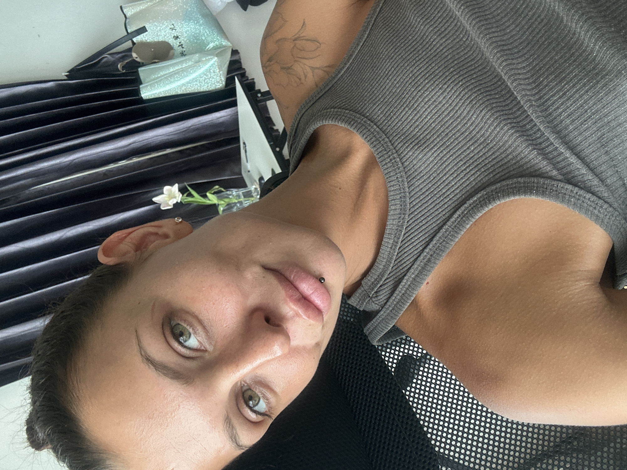 A young woman with light brown skin and green eyes is photographed in a close-up, selfie-style shot. She has her dark hair pulled up in a bun. She is wearing a gray ribbed tank top. A lip piercing is visible on her lower lip. A floral tattoo is visible on her right shoulder. The background appears to be an indoor setting with dark curtains and a white table. A sparkly tote bag is hanging on the wall in the background. The image was taken in Ko Samui District, Thailand.