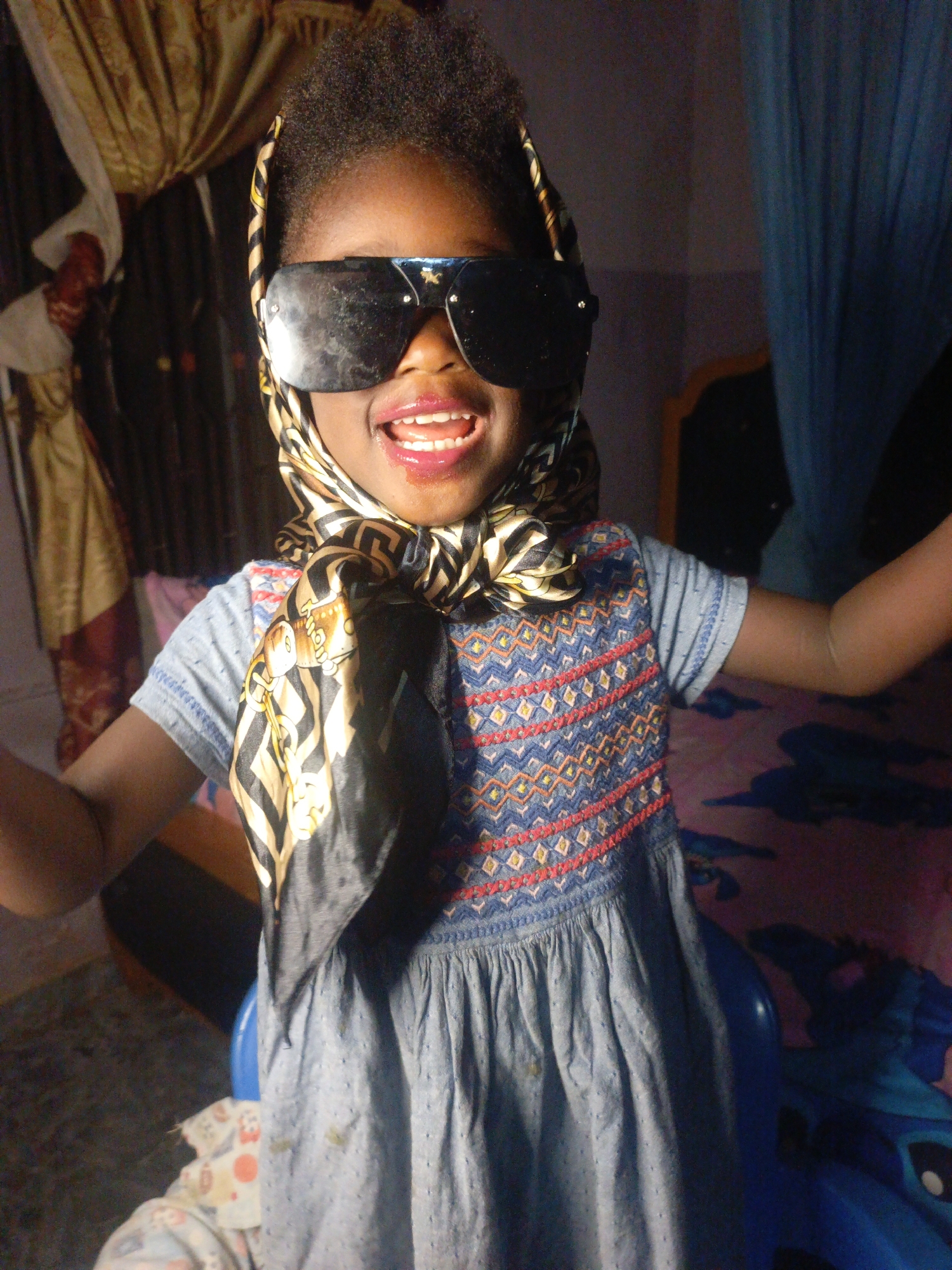 A young child stands indoors in Akwanga, Nigeria. The child wears a light blue dress with geometric embroidery on the upper part, oversized black sunglasses, and a patterned scarf tied around their head. The child's mouth is open, possibly smiling.
Behind the child, there is a draped golden curtain and a bed with a blue blanket featuring cartoon characters. A partially visible painting or print hangs on the wall behind the curtain. A mosquito net is draped to the right of the frame.