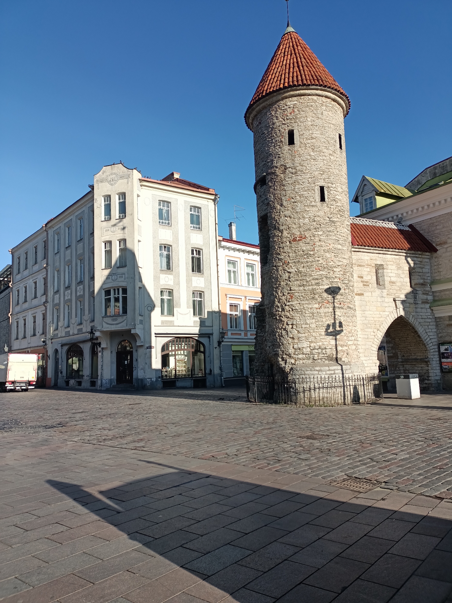 The image shows a street scene in Tallinn, Estonia, featuring a historical tower and surrounding buildings. On the right is a round stone tower with a red tiled conical roof, characteristic of medieval architecture. To its left is a large arched gateway made of similar stone, suggesting it is part of a fortification or an old city entrance. Adjacent to the tower and gateway are several multi-story buildings with light-colored facades and numerous windows. One building in the foreground has a distinct shadow cast on its facade, shaped like a pointed arch. The street in front of the buildings is paved with cobblestones, and a long shadow from an unseen object stretches across the foreground. A white truck is parked on the left side of the street. The sky is clear and blue, indicating a sunny day. The overall scene depicts a well-preserved historical urban environment.