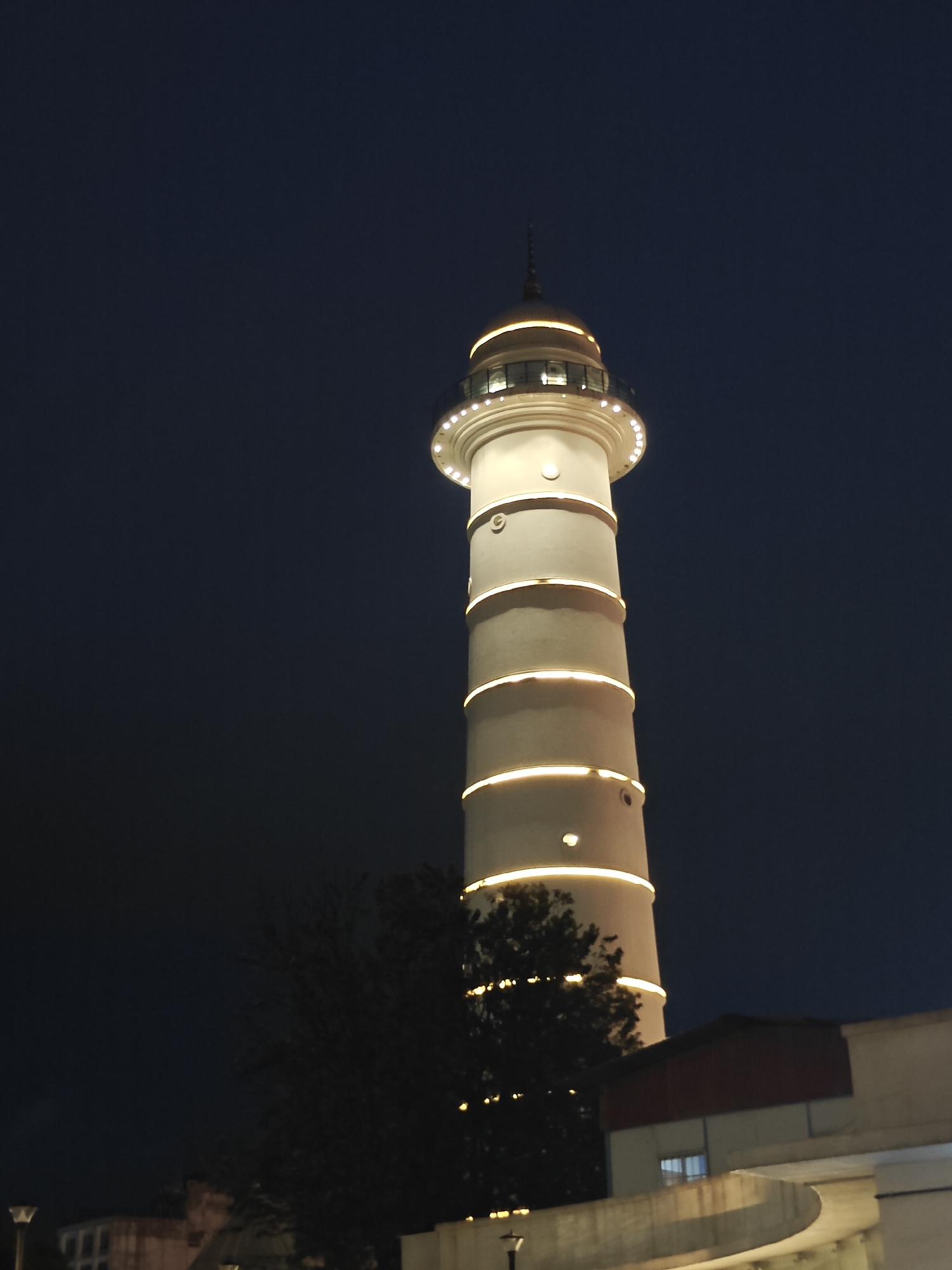 A white, cylindrical tower illuminated by horizontal bands of yellow light stands against a dark blue night sky. The tower has a golden dome at its peak, topped with a spire. Circular windows are visible at various levels of the tower, and a ring of lights encircles the base of the dome. In front of the tower, a dark, leafy tree partially obscures the lower section. To the right of the tree, a building with a red roof and windows is visible. The foreground shows a curved concrete structure. The scene is located in Kathmandu Metropolitan City, Nepal.