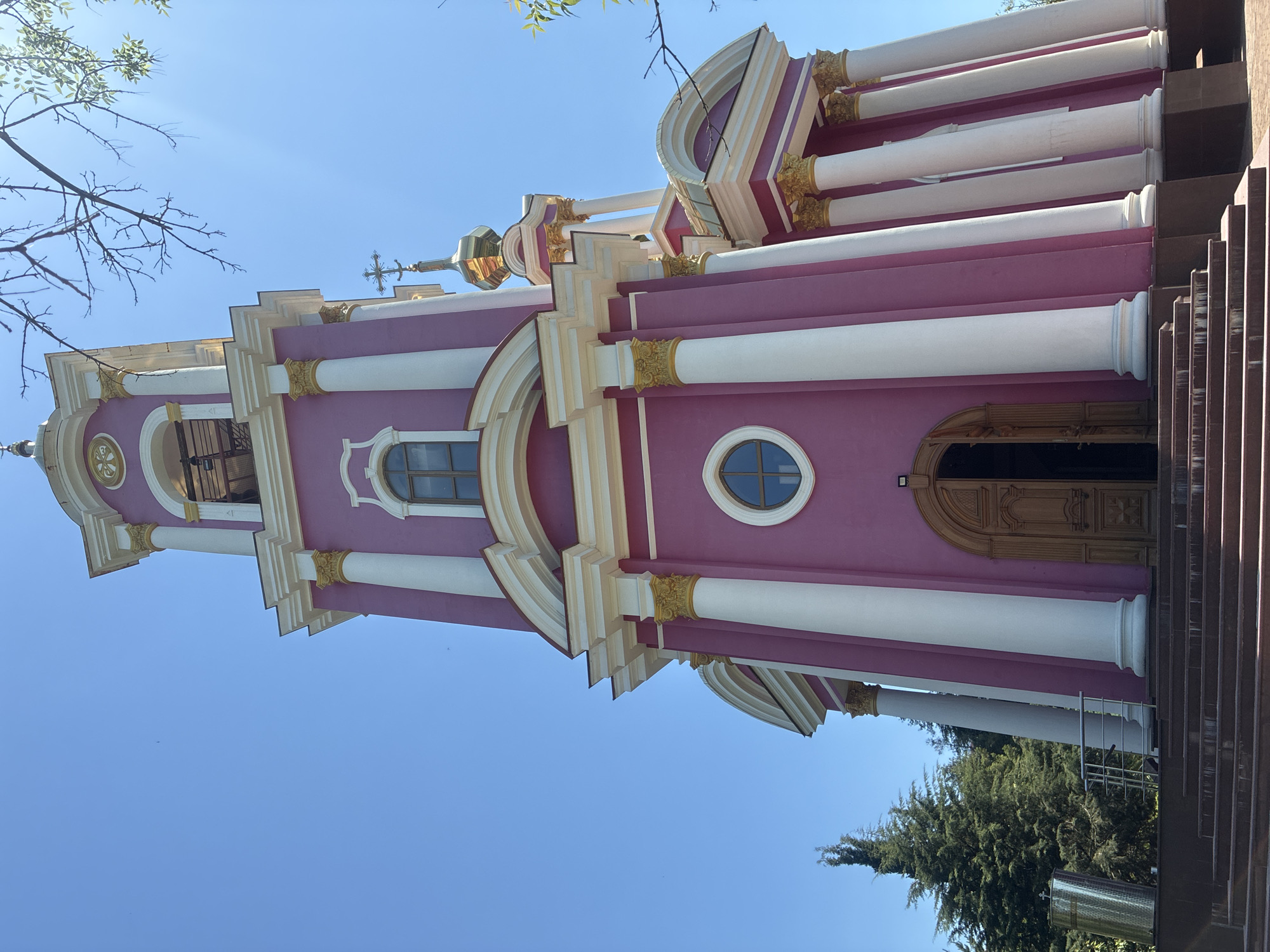 The image displays an ornate church building and its bell tower under a clear, bright blue sky in Sochi, Russia. The architecture is characterized by vibrant magenta-pink walls accented with numerous white columns, arched details, and cornices. Gold gilding is prominent on the column capitals, decorative friezes, and the domes and finials topping the structure.
A tall bell tower rises on the left, featuring multiple levels with open archways and topped by a golden dome and a cross. The main facade of the church incorporates vertical white columns, many with intricate golden Corinthian-style capitals. Windows are a mix of arched and circular shapes, with visible glass panes. Towards the right, a large, double wooden door with detailed carving serves as the main entrance. A wide set of dark, multi-tiered stairs leads up to this entrance.
The scene is set outdoors on a sunny day, indicated by the clear blue sky and bright illumination. Tree branches with green leaves are visible in the upper left corner, and a dense evergreen tree stands near the base of the building on the right. A silver, cylindrical metal tank is partially visible on the ground next to the tree. No people or discernible text are present in the image. The overall impression is one of a richly decorated and well-maintained religious edifice.