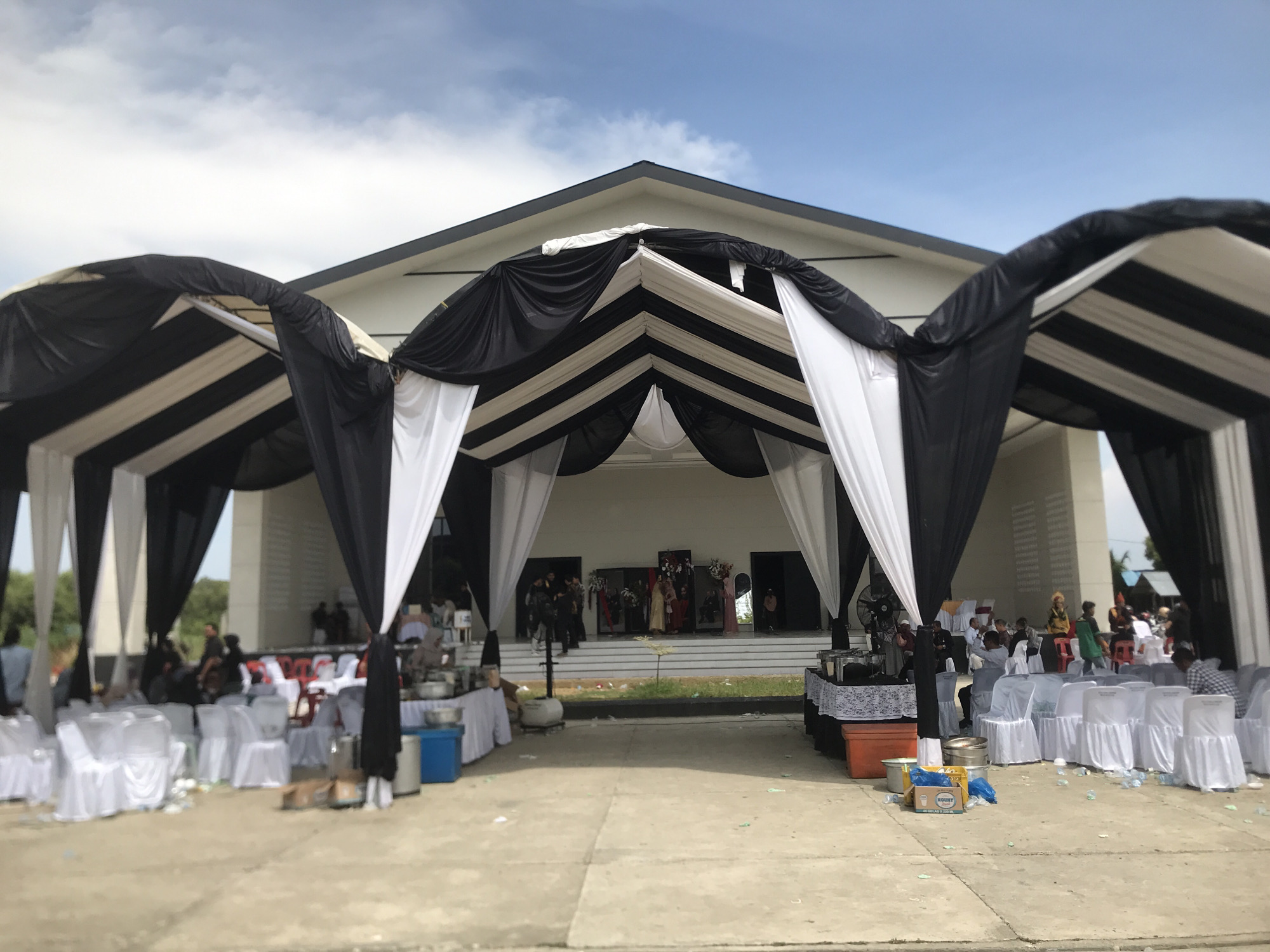 The image captures an event taking place in Lhokseumawe, Indonesia. A building with a peaked roof serves as the backdrop. An awning of alternating black and white fabric draped over a frame extends from the building, creating a covered area. Numerous white chairs are arranged under the awning, some occupied by people. Tables, some with food and beverage containers, are also present under the awning. In the background, people are seen on the steps of the building. The environment appears to be outdoors, with a concrete ground surface. The sky is visible above the building.