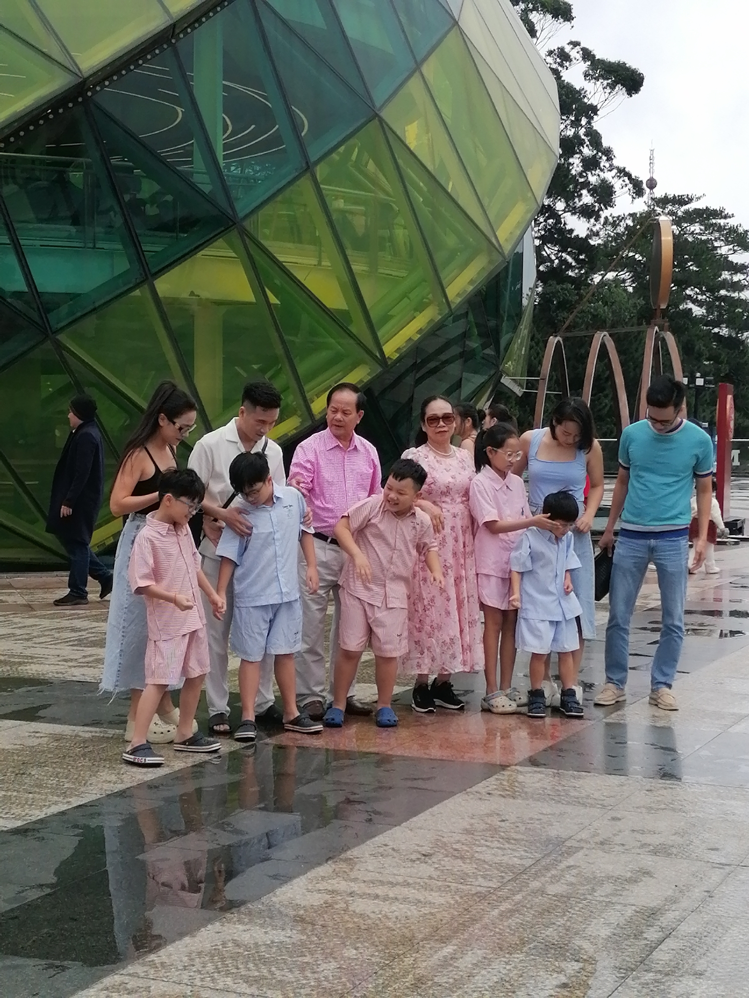 The image depicts a group of people standing in front of a large, geometric, glass-paneled building with a green and yellow hue. The building's design is multifaceted and contemporary. In the foreground, a family group consisting of adults and children is posing for a photograph. The adults appear to be two males and two females, while there are at least three children visible, two boys wearing light blue outfits and one boy and one girl in pink and white striped outfits. The children are positioned closer to the camera, with the adults behind them. A man in a pink checkered shirt stands to the right of a woman in a floral dress, who is in turn next to a young girl. To the left of the woman in the floral dress is a boy in a pink striped outfit. Further to the left, a man in a white shirt stands with his arm around a boy in a blue outfit. A woman in a black tank top and a long denim skirt is to the left of the man in the white shirt, holding hands with a boy in a pink striped outfit. The ground in front of the group is wet, suggesting recent rain, and reflects the building and the sky. In the background, trees and part of a metallic sculpture are visible. The overall setting appears to be an outdoor public space. This scene is consistent with the exterior of the Da Lat Flower Garden attraction in Da Lat, Vietnam, known for its modern architectural elements and landscaped grounds.