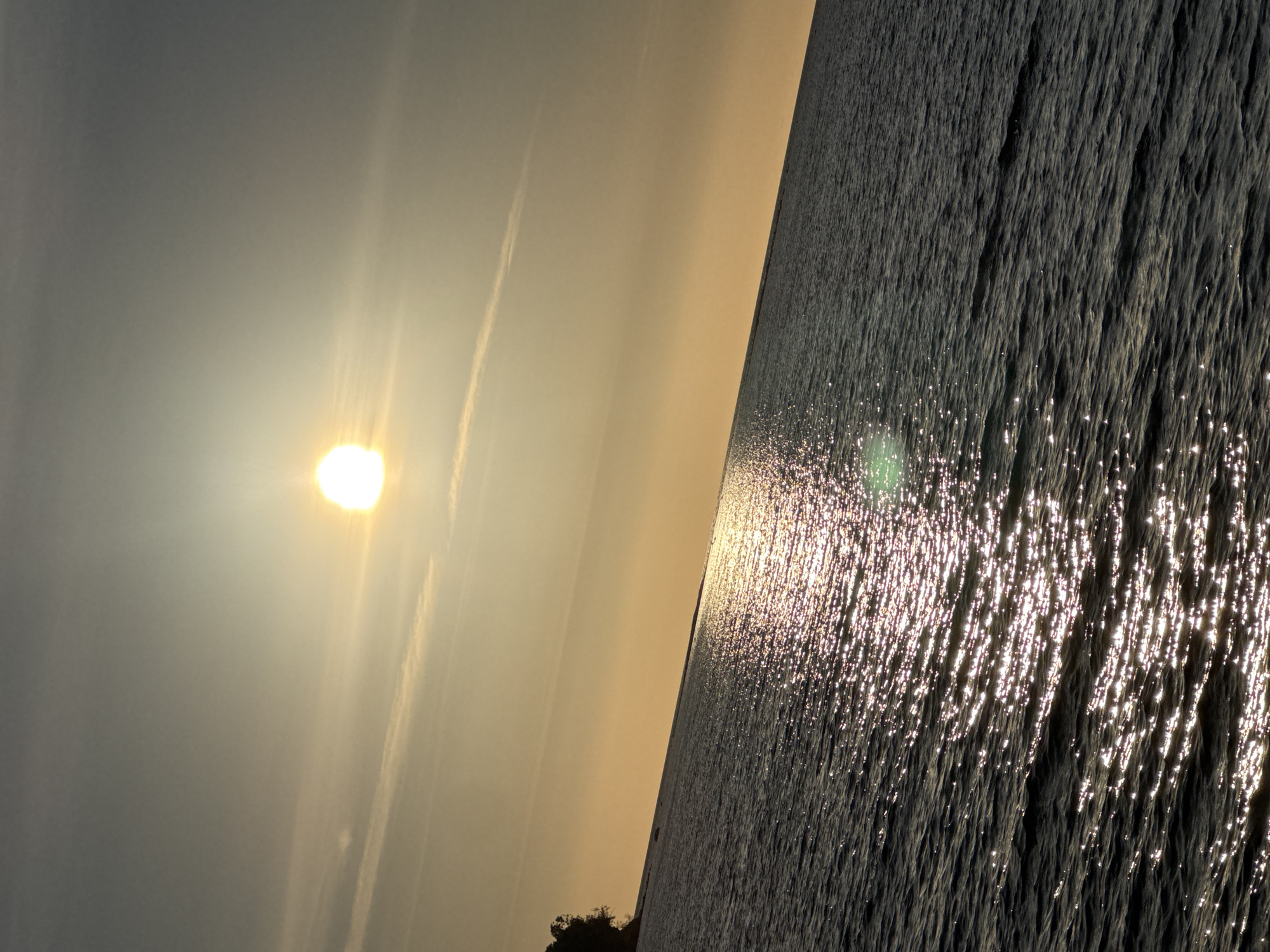 The image shows a sunset over the sea. The sun is a bright orb in the sky, casting a golden glow over the water. The sea is dark and choppy, with waves reflecting the sunlight. There is a small island or landmass visible in the distance on the left side of the image. The sky is clear with a few wispy clouds. The scene is a natural landscape, likely a coastal area. Based on the visual cues, such as the type of coastline and the typical sunset conditions, this scene could be indicative of the Baltic Sea coast near Tallinn, Estonia, possibly from the Pirita beach or the coastline further north or west.