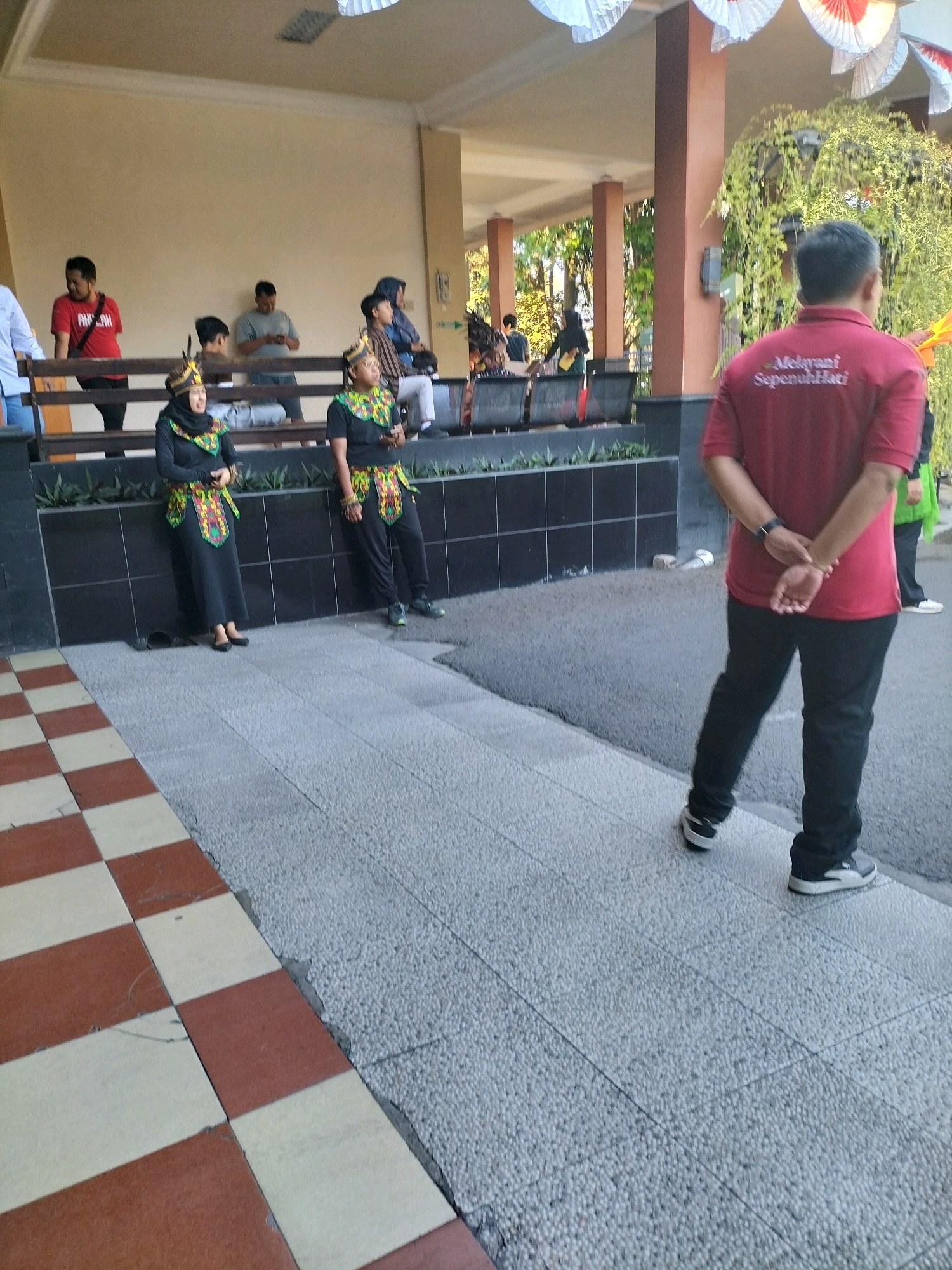 The image depicts an outdoor scene in front of a building, possibly a school or community center, in the city of Pare, Indonesia. In the foreground, a man wearing a red polo shirt with text that appears to read "Melayani Sepenuh Hati" (Serve with All Your Heart) stands with his back to the camera, facing away from the building. He is wearing black pants and athletic shoes. Two women, dressed in traditional attire that includes black headscarves and colorful patterned vests over black outfits, stand to the left of the man, closer to the building. The woman on the left is wearing a long black dress, while the woman on the right is wearing black pants. Several other individuals are visible in the background, some standing and some sitting on a bench. The building has an open-air corridor with columns and decorative white and red paper elements hanging from the ceiling. Lush green foliage is visible on the right side of the building. The ground is a mixture of patterned tiles and textured concrete.