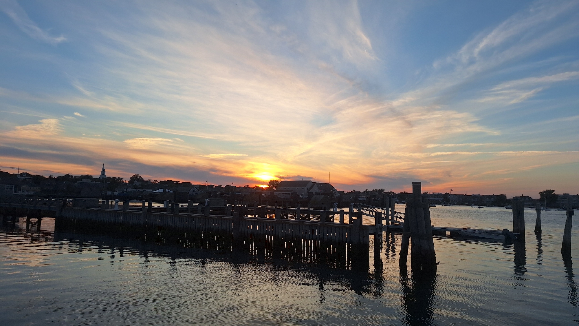 This is a horizontal, eye-level, full shot of a sunset over a harbor in Nantucket, Massachusetts. The sky is a gradient of blue, with streaks of orange, yellow, and pink from the setting sun. Wispy clouds stretch across the sky, reflecting the colors of the sunset. In the foreground, a wooden dock extends into the water, with its pilings casting dark reflections on the rippling surface. The water is a dark, glassy blue, mirroring the sky and dock. In the background, a small town with white buildings and a steeple is visible along the shoreline. Several boats are docked in the harbor. The scene evokes a sense of tranquility and natural beauty.