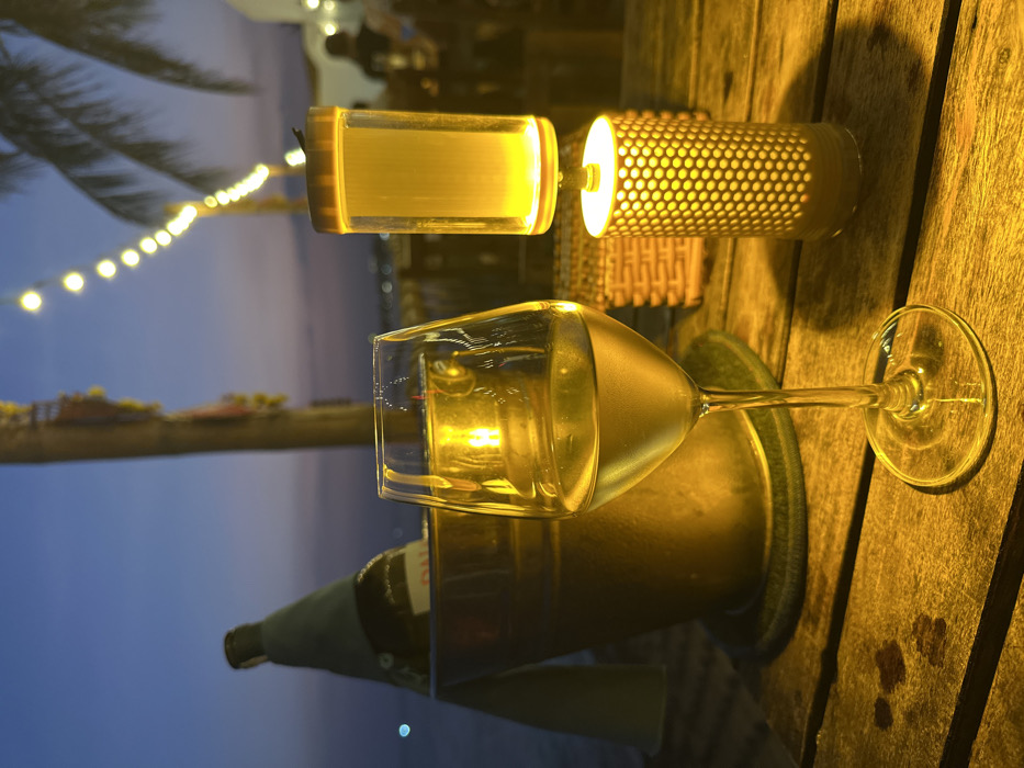 The image depicts an outdoor evening scene at a waterfront establishment in Thành phố Phan Thiết, Vietnam, captured from a low perspective looking across a wooden slatted table. In the immediate foreground, a clear wine glass, approximately half-filled with a pale yellow liquid (likely white wine), stands prominently. To its right, a two-part decorative lamp emits a warm yellow light; its lower section is a cylindrical lantern with a perforated, honeycomb-like pattern, while the upper section is a clear, cylindrical container. To the left and slightly behind the wine glass, a metallic ice bucket holds a dark-colored bottle, partially covered by a light-colored fabric or sleeve, resting on a dark coaster. A small, dark-colored woven basket is visible on the table behind the lamp.
The background reveals a twilight sky, transitioning from deep blue at the top to lighter purplish-orange hues near the horizon line. A body of water is visible to the left, showing faint reflections or distant lights. A string of warm white globe lights extends diagonally across the mid-ground. Silhouetted palm tree fronds are visible on the right. In the far background, blurred figures of people and illuminated structures (likely part of a restaurant or bar) are discernible, along with additional distant lights on the horizon. The overall environment suggests an ambient, relaxed social setting.
