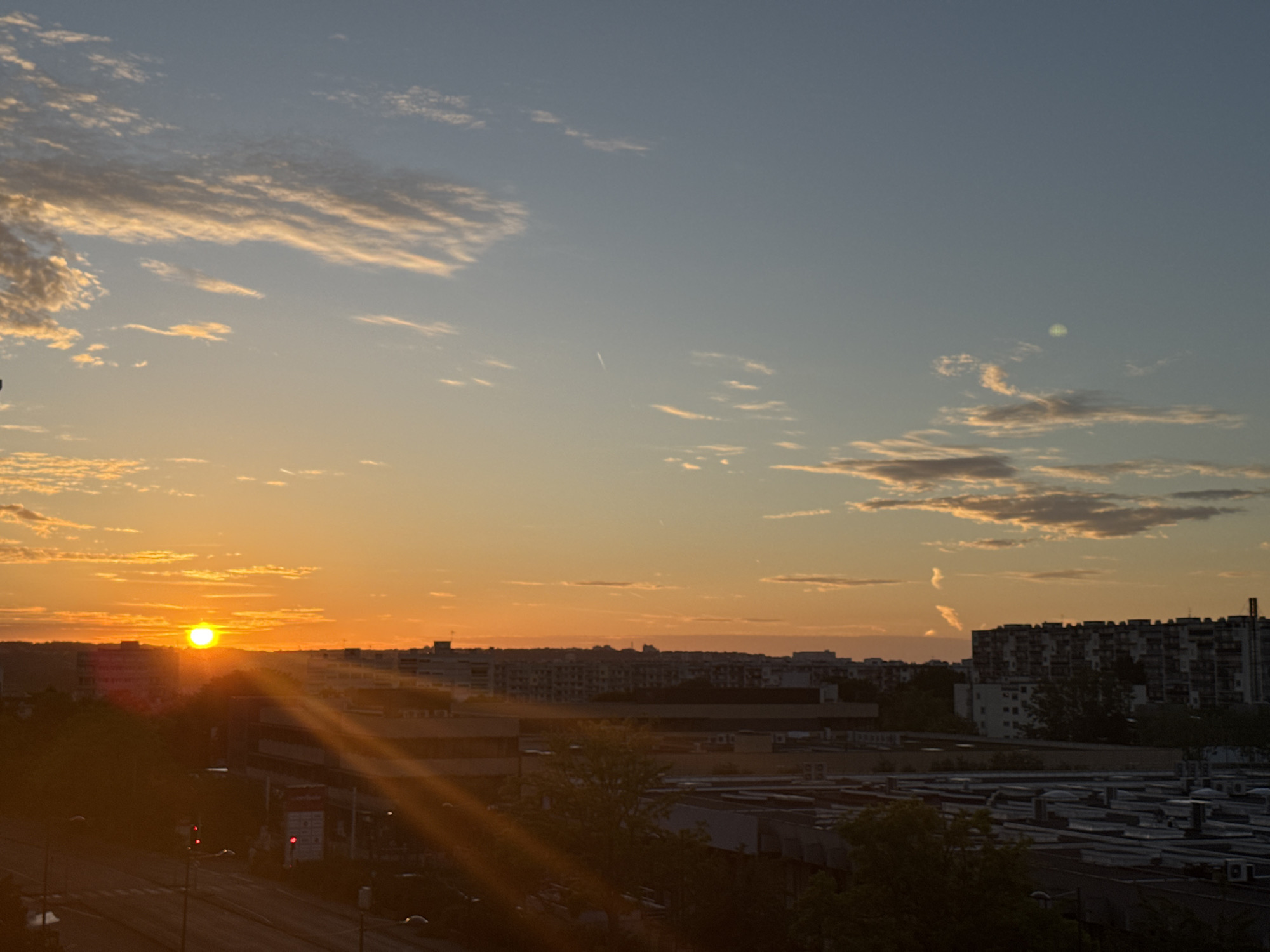 This is an eye-level, wide-angle photograph of a city skyline at sunset. The sun is a bright orb setting on the left side of the image, casting long rays of light across the sky and over the buildings. The sky is a gradient of orange and blue, with scattered clouds that are illuminated by the setting sun. In the foreground, there is a road with traffic lights, and a street with streetlights. In the middle ground, there are several buildings of various sizes, including apartment buildings and industrial buildings. The scene is one of a city transitioning from day to night.