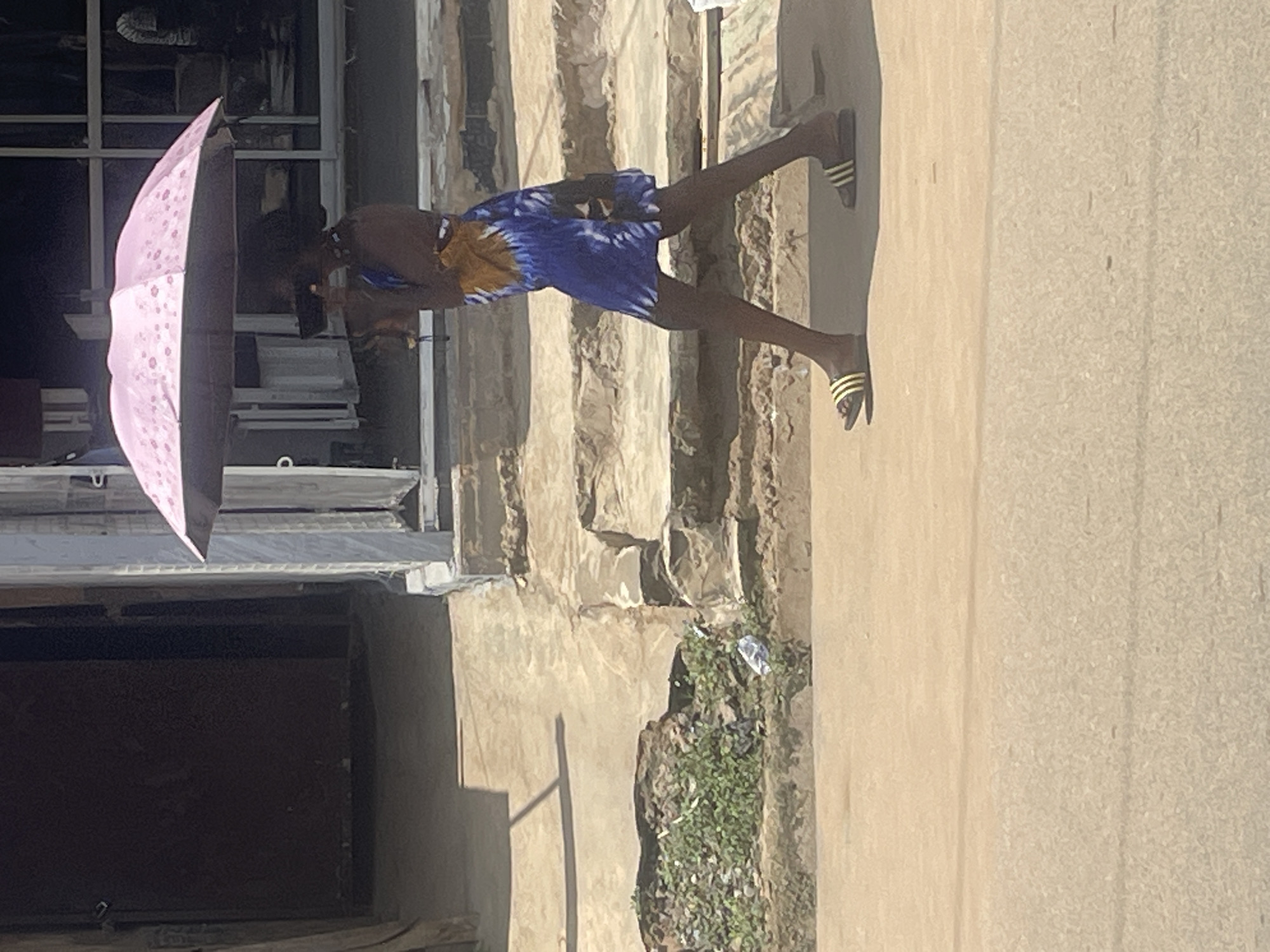 A full-body, side-view shot depicts a person with dark skin walking along a paved path in Abuja, Nigeria. The individual is holding a pink umbrella overhead. They are wearing a blue and yellow patterned dress and dark sandals with yellow stripes. The environment includes a building with large windows on the right and a darker structure on the left. A patch of green vegetation with some debris is visible to the left of the paved path. The ground in the foreground is a light brown, sandy color, suggesting an outdoor, possibly sunlit, setting.