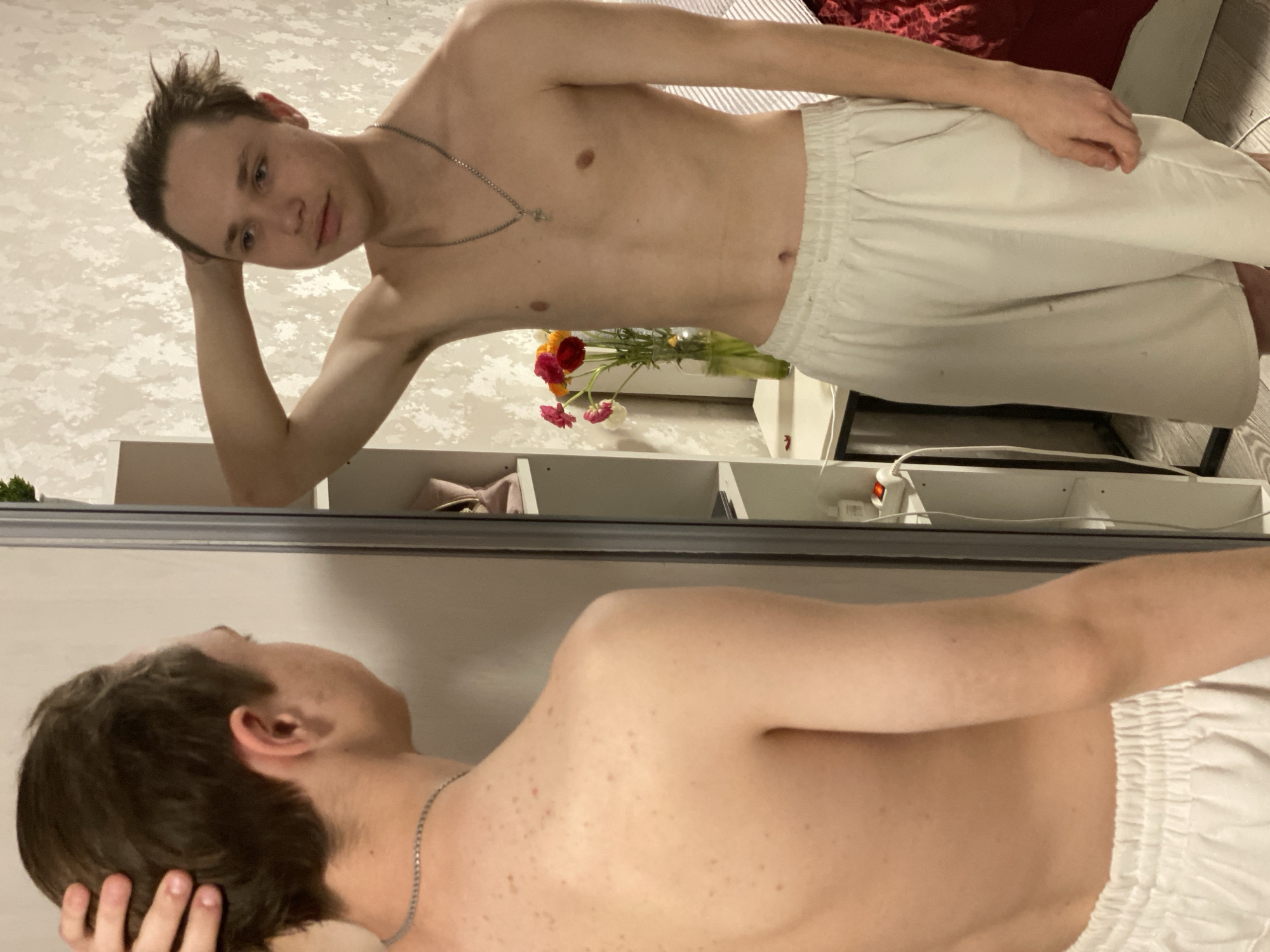 This image captures a young, lean individual, likely male, posing shirtless in an indoor setting. He has short, dark hair styled upwards on top and fair skin. He is wearing light-colored (possibly white or off-white) elastic-waist shorts and a thin silver chain necklace with a small, indistinct pendant. His left arm is bent, with his left hand resting on the side of his head or ear. He looks directly at the camera with a neutral or slightly relaxed expression. The image itself appears rotated 90 degrees counter-clockwise.
The background features a light-colored wall with a subtle patterned texture. Behind the subject's torso, a small vase holds a colorful bouquet of flowers, including red, orange, white, and pink blossoms, placed on a light-colored surface. To the right and further back, a glimpse of dark red, textured fabric, possibly a bedspread or couch, is visible.
The lower portion of the image is dominated by a large mirror, reflecting the back of the individual's head, right shoulder, and arm. Within this reflection, light-colored shelving units or furniture, along with an electrical outlet and a white cord, can be seen. The overall setting suggests a casual personal space, such as a bedroom. The lighting is soft and even, indicating an indoor environment. There is no visible text in the image. This scene is situated in Sochi, Russia.