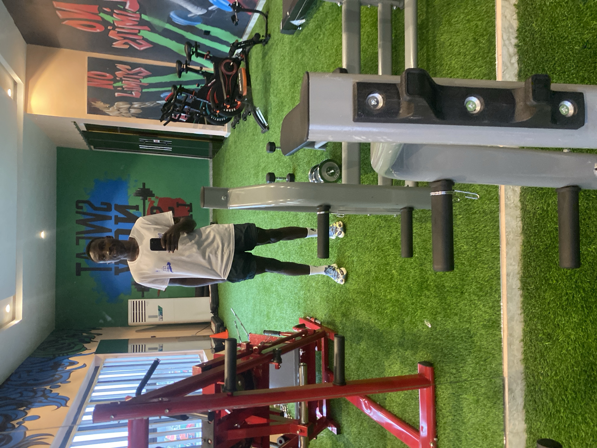 A man stands in a brightly lit gym, holding a smartphone and taking a selfie. He is wearing a white t-shirt, dark shorts, and athletic shoes. The gym features artificial turf flooring and walls decorated with motivational graphics, including the words "SWEAT NOW" and "NO LIMITS." Exercise equipment, such as stationary bikes and weight machines, is visible in the background. The gym appears to be located in Barden Yana, Nigeria.