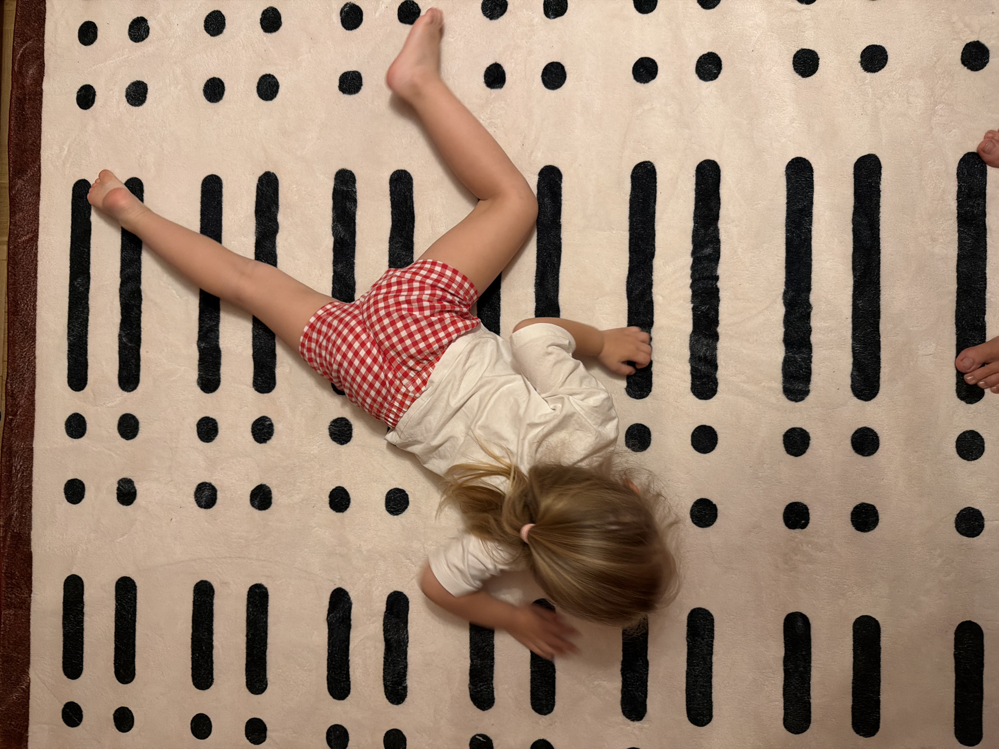 A high-angle shot shows a young girl lying on a patterned rug. The rug is light-colored with black spots and vertical lines in a repeating pattern. The girl is wearing red and white checkered shorts and a white shirt. Her legs are splayed out to the left, and she is lying on her stomach. Her head is near the bottom of the frame, and her hair is tied back in a ponytail. The location is unspecified and outside the scope of identification as Khimki, Russia.