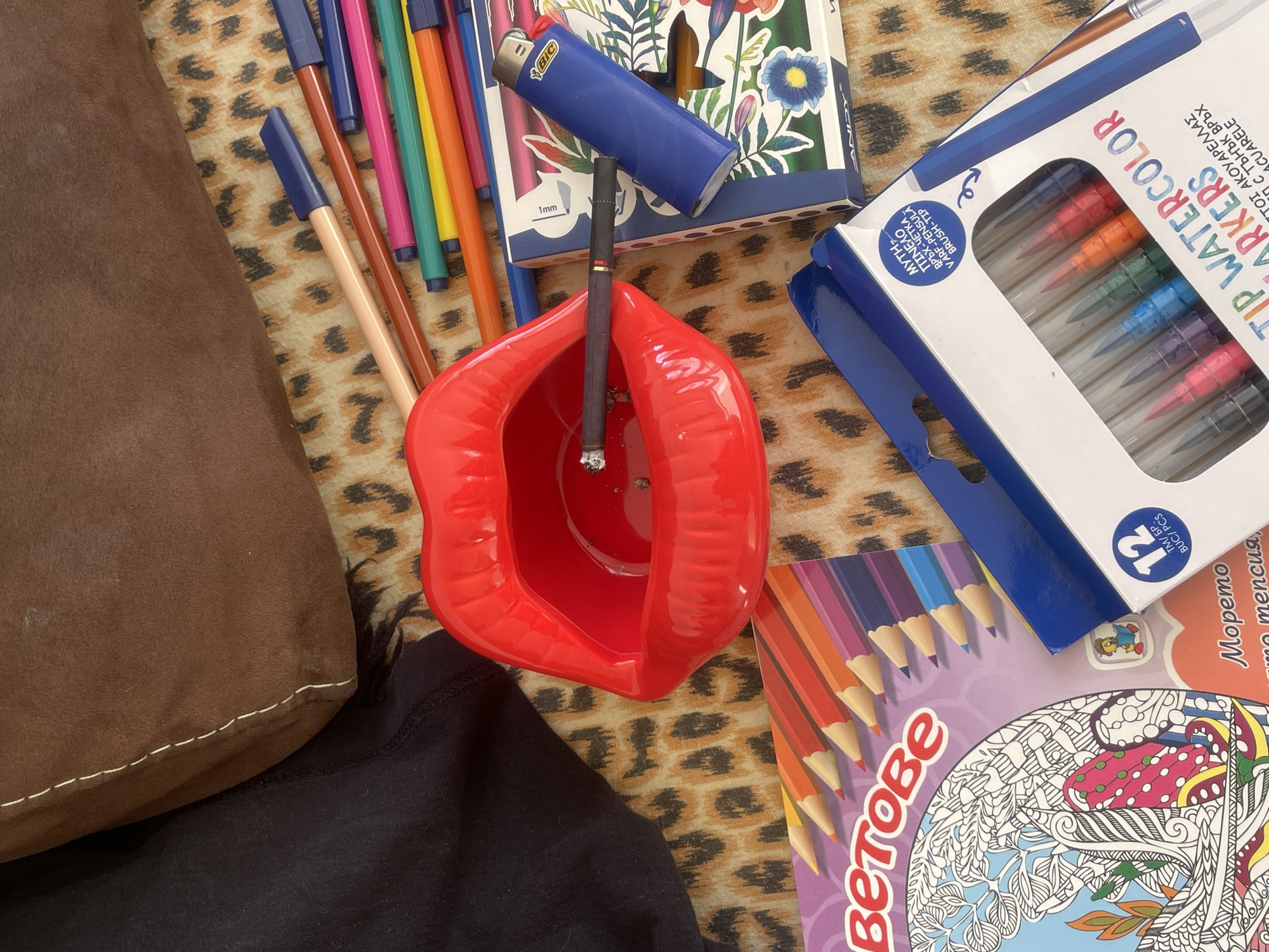The image depicts a close-up, overhead view of a collection of art supplies and smoking paraphernalia laid out on a leopard-print fabric surface, likely a throw or blanket. A bright red, lip-shaped ashtray is prominently featured, containing a lit cigarette resting on its edge. Beside the ashtray are several blue and black colored pencils, a blue Bic lighter, and a package of watercolor markers. A coloring book with an intricate design of trees and mushrooms is also visible. The scene is lit by natural light, casting soft shadows. There is no indication of a specific location within the city of Varna, Bulgaria.