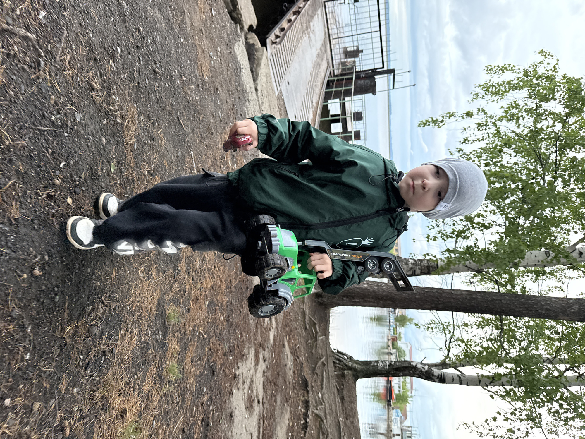 A child stands outdoors near the shoreline of the Kama River in Chaikovsky, Russia. He is wearing a grey beanie, a dark green jacket, and dark trousers with white text on the side. He is holding a green plastic toy truck. In the background, there is a pier with metal railings, the Kama River, a cloudy sky, and birch trees. The ground is covered with dirt, grass, and dried leaves.