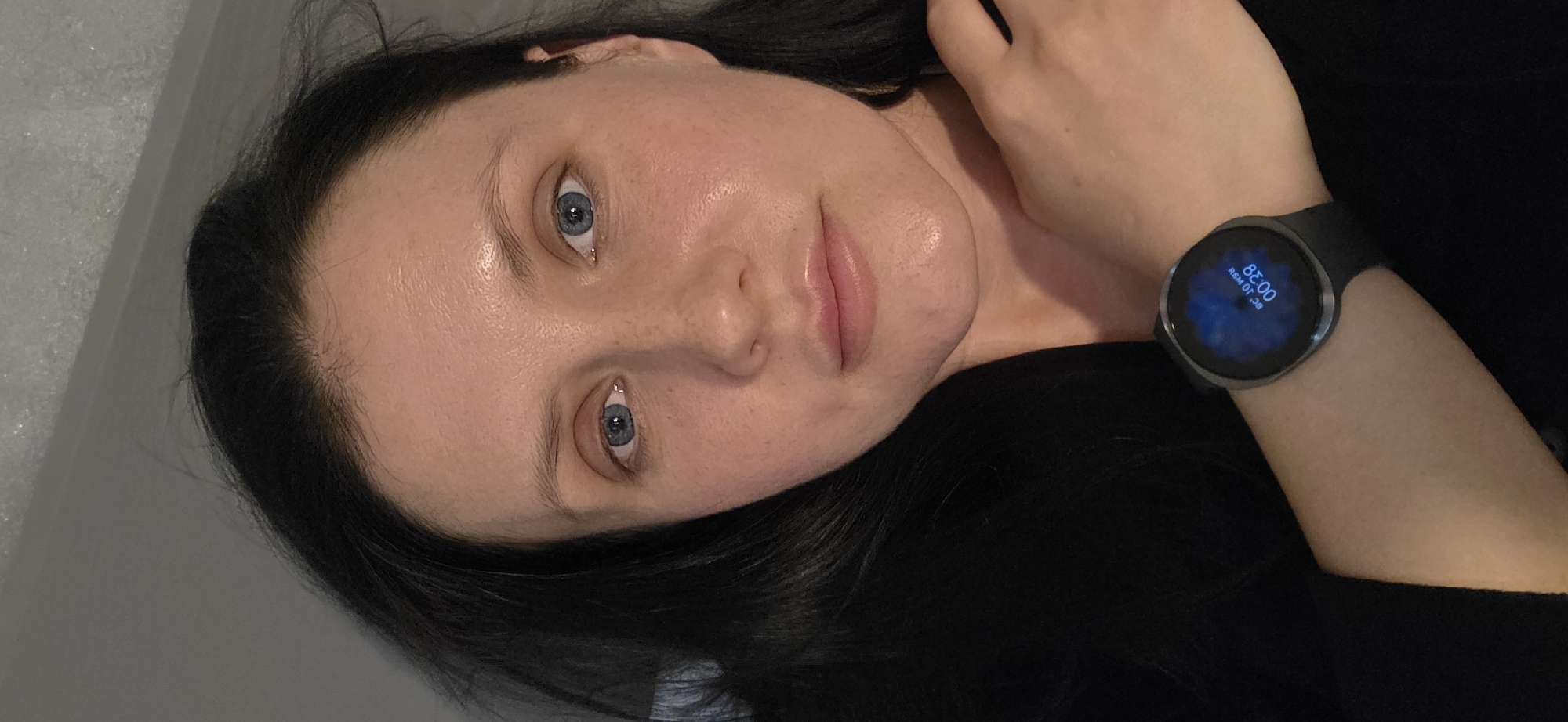 A young woman with fair skin, dark hair, and blue eyes is positioned at an angle in the photograph. She is wearing a dark-colored top. On her left wrist is a smartwatch displaying a blue screen with the time "00:38" and the date "10 OKT." Her right hand is near her neck. The background is a light grey or white wall. The location is not identifiable as Spijkenisse, Netherlands from the image alone.