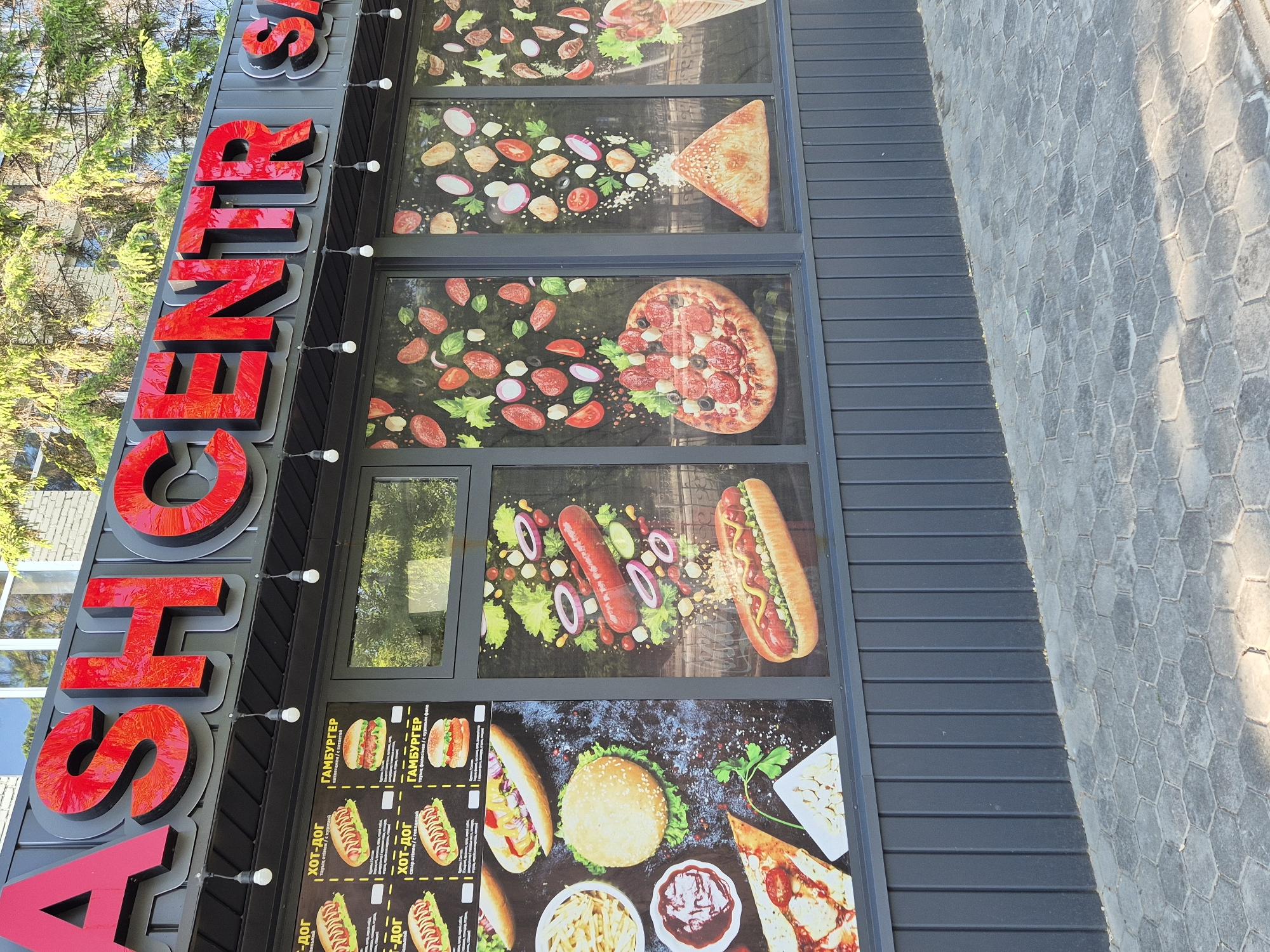Here's a concise description of the image:
The image shows the exterior of a food establishment, likely a fast-food outlet, identified by the large red text "ASH CENTER" running vertically on the left side. The building's exterior is dark grey with what appears to be a black, grooved or paneled facade. The windows are covered with promotional images of food items, including pizza, hot dogs, and burgers. There are menus visible with Cyrillic text, suggesting the location is in a Russian-speaking region. The text visible mentions "HOT-DOG" and "HAMBURGER" in Cyrillic. The ground adjacent to the building is paved with hexagonal tiles.