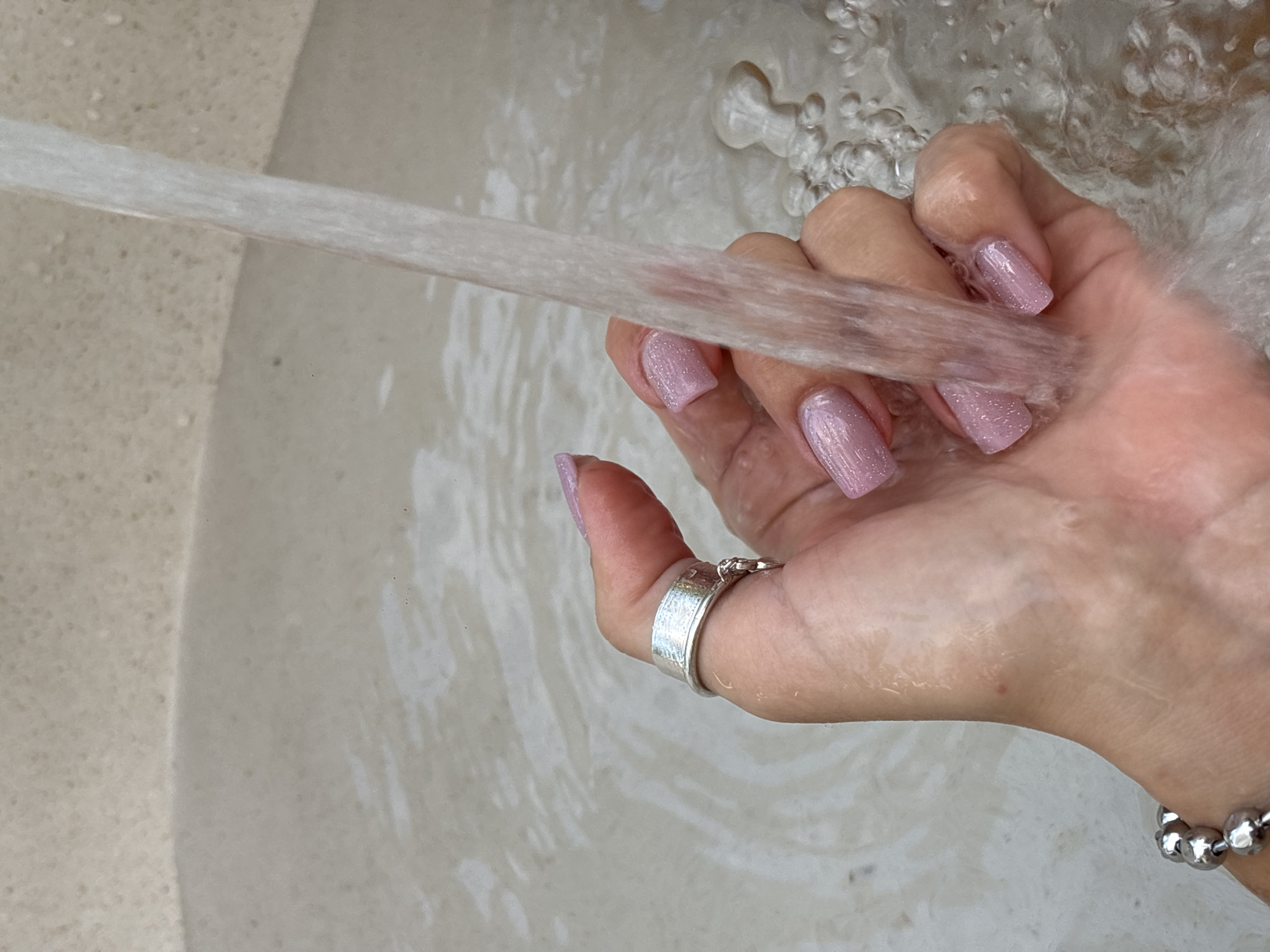 An overhead, close-up shot captures a person's hand and forearm submerged in a light-colored, textured sink filled with running water. The hand is positioned to the left of the frame, with the forearm extending towards the bottom left. The water flows from the upper right corner, cascading over the hand and creating ripples and bubbles in the sink. The person's nails are painted a sparkling light pink, and they wear a silver ring on their index finger and a silver beaded bracelet on their wrist. The sink appears to be made of a speckled material, possibly granite or quartz.