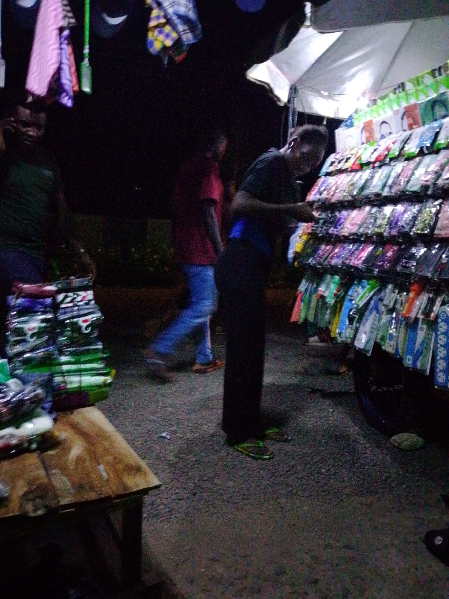 The image depicts a nighttime street scene in Jalingo, Nigeria, likely at a market stall. In the foreground, a wooden table is visible, laden with stacks of plastic-wrapped items, possibly phone cases or accessories. To the right, a person wearing a black t-shirt and dark pants is browsing a stall displaying a variety of colorful phone cases. The stall is illuminated by an overhead light, and a white umbrella provides shade. In the background, another person in a red shirt and blue jeans is walking away from the camera. Above the stall, several pieces of clothing are hanging, including a pink patterned garment and a blue and yellow checkered fabric. The ground is asphalt, suggesting an urban outdoor setting. The overall impression is that of a busy market or street vendor operation at night.