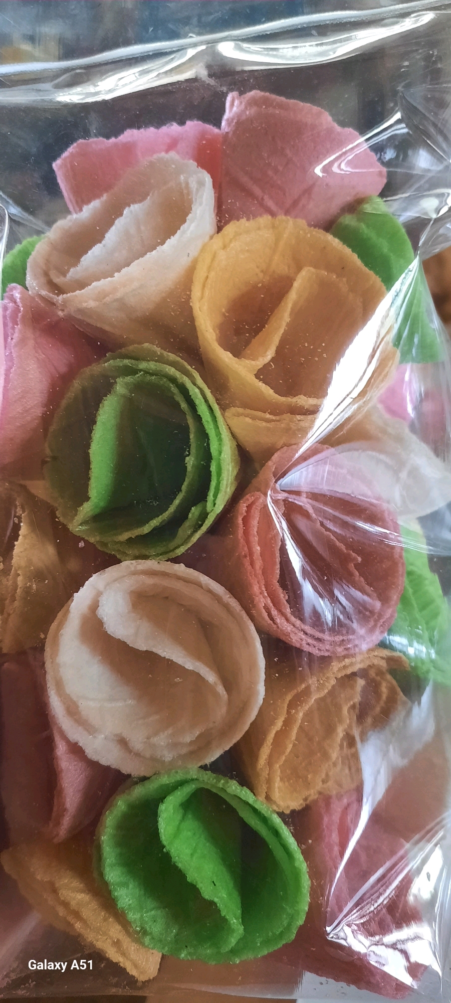 A clear plastic bag contains a close-up, vertically oriented view of colorful, rolled snacks. The snacks, which appear to be made of a crisp, wafer-like material, are arranged in a dense cluster. They exhibit a variety of pastel colors including pink, light cream, yellow, green, and reddish-orange. The rolled shape of each snack resembles a coiled petal or a rosebud. The texture of the snacks appears slightly rough or granulated. Reflections from the plastic packaging are visible on the surface of the snacks, obscuring some details and adding a glossy sheen to the image. The background is dark and out of focus, drawing attention to the brightly colored snacks. The bottom left corner of the image displays white text that reads "Galaxy A51". The image is not specific to any location in Ploso, Indonesia.