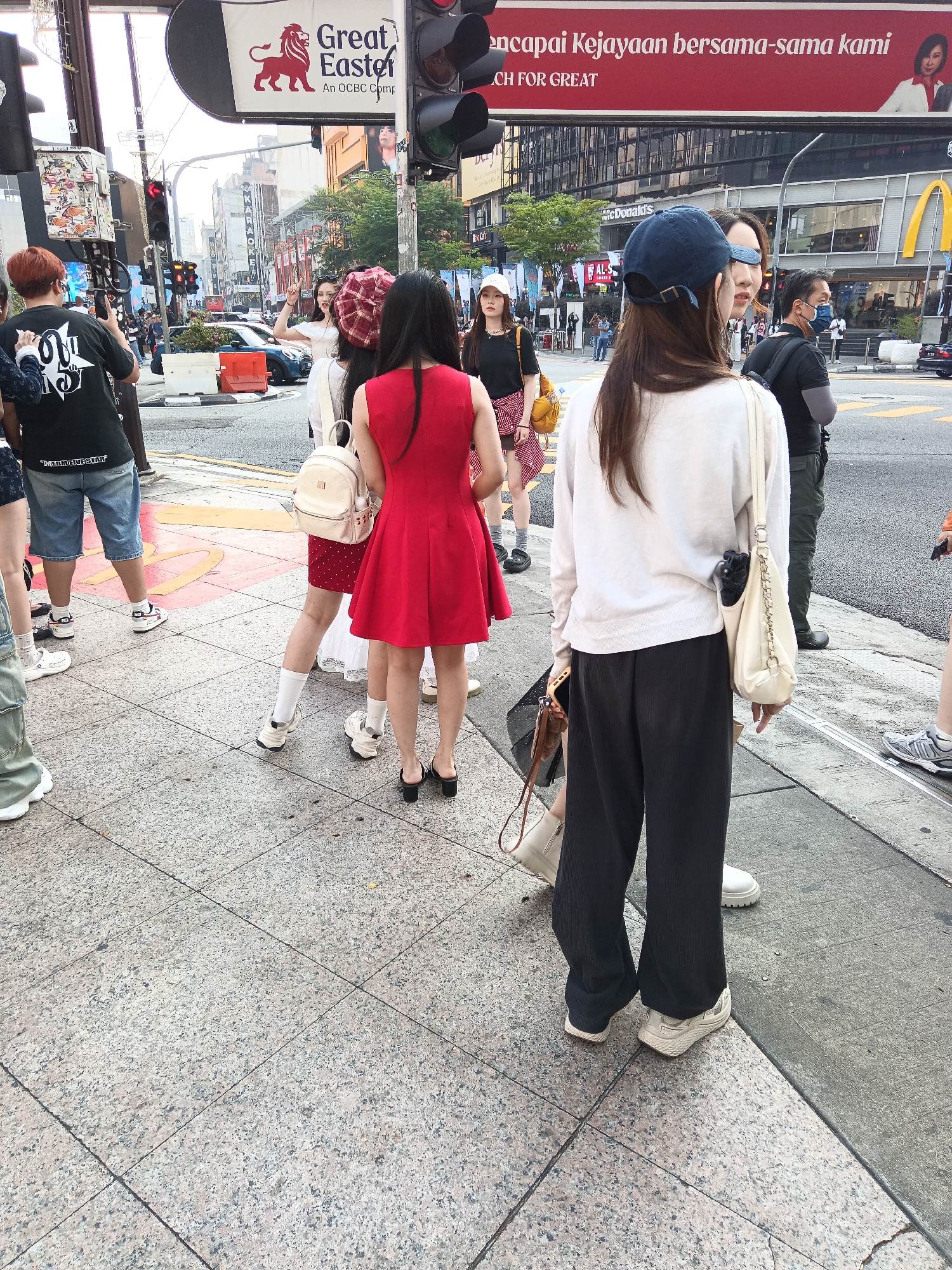 The image captures a street scene in Kuala Lumpur, Malaysia, likely in the busy Bukit Bintang area. In the foreground, several individuals are standing on a pedestrian walkway next to a street. A woman in a red dress and a plaid headscarf is the most prominent figure, facing away from the camera. To her left, a person with red hair is holding up a phone, appearing to take a photo or video. Other people are visible, including a woman wearing a blue baseball cap and a white long-sleeved shirt, and a man in a black t-shirt and a blue face mask. Behind them, a McDonald's restaurant with its iconic golden arches is identifiable, suggesting a commercial district. Traffic lights and street signs are visible in the background, indicating an urban intersection. The pavement is made of granite tiles.