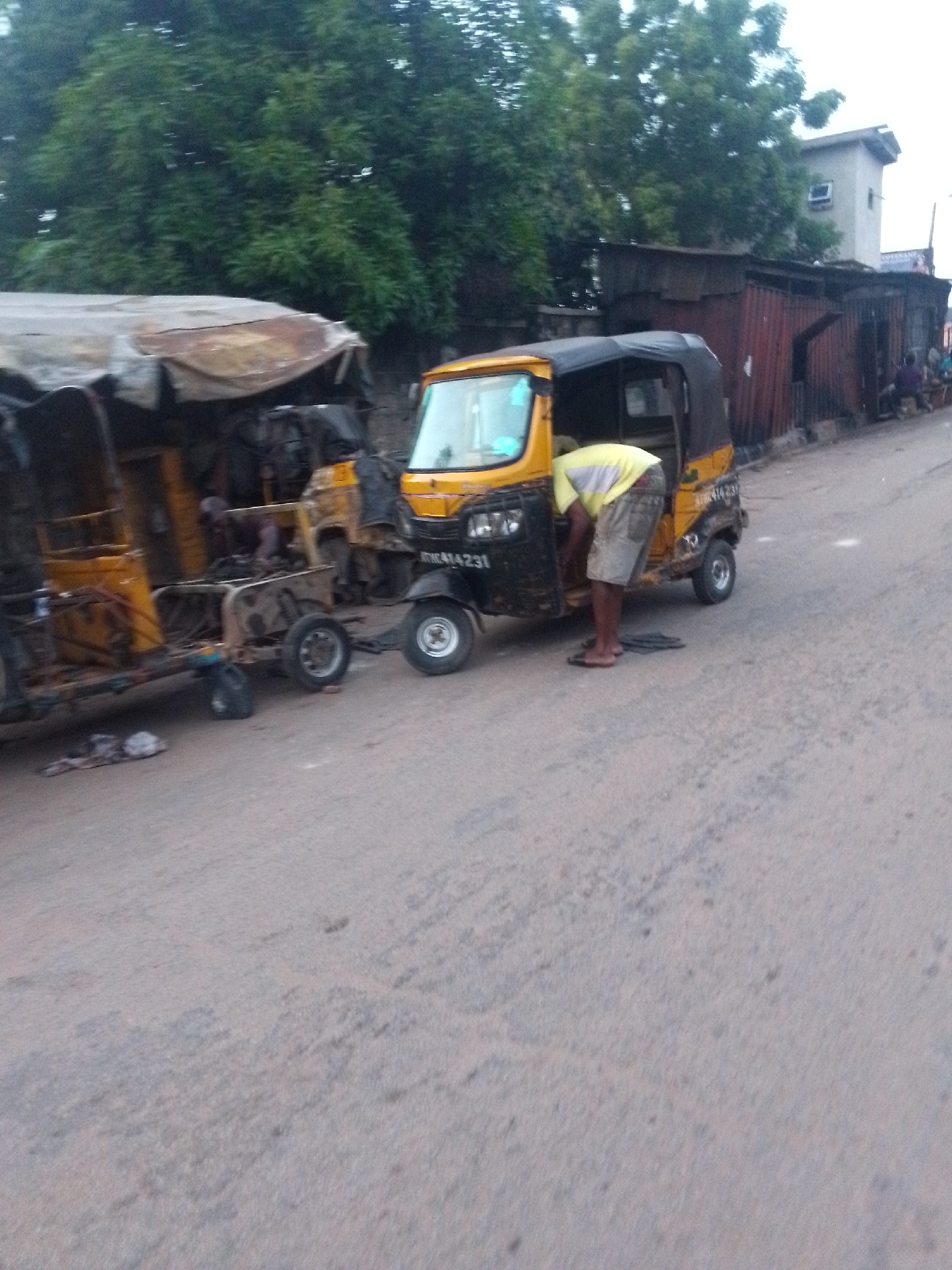 A person is working on a yellow tricycle on a city street in Unknown, Unknown. The tricycle appears to be damaged. There is a damaged tricycle towed behind the first one. The person is wearing a light-colored shirt and shorts. In the background are trees, buildings, and other people. The street is paved but in disrepair.