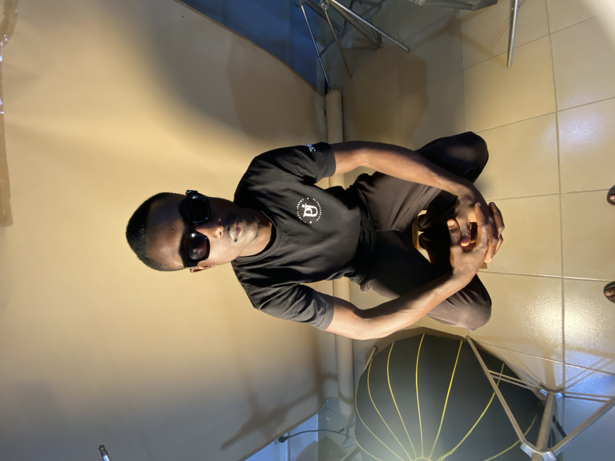 A dark-skinned man wearing sunglasses and a black t-shirt with a logo kneels on a tiled floor. He is positioned in the center of the frame, facing forward with his hands clasped in front of him. The background consists of a light-colored, textured wall and photographic equipment, including a large black umbrella-shaped light modifier with yellow lines. The lighting suggests an indoor studio setting.