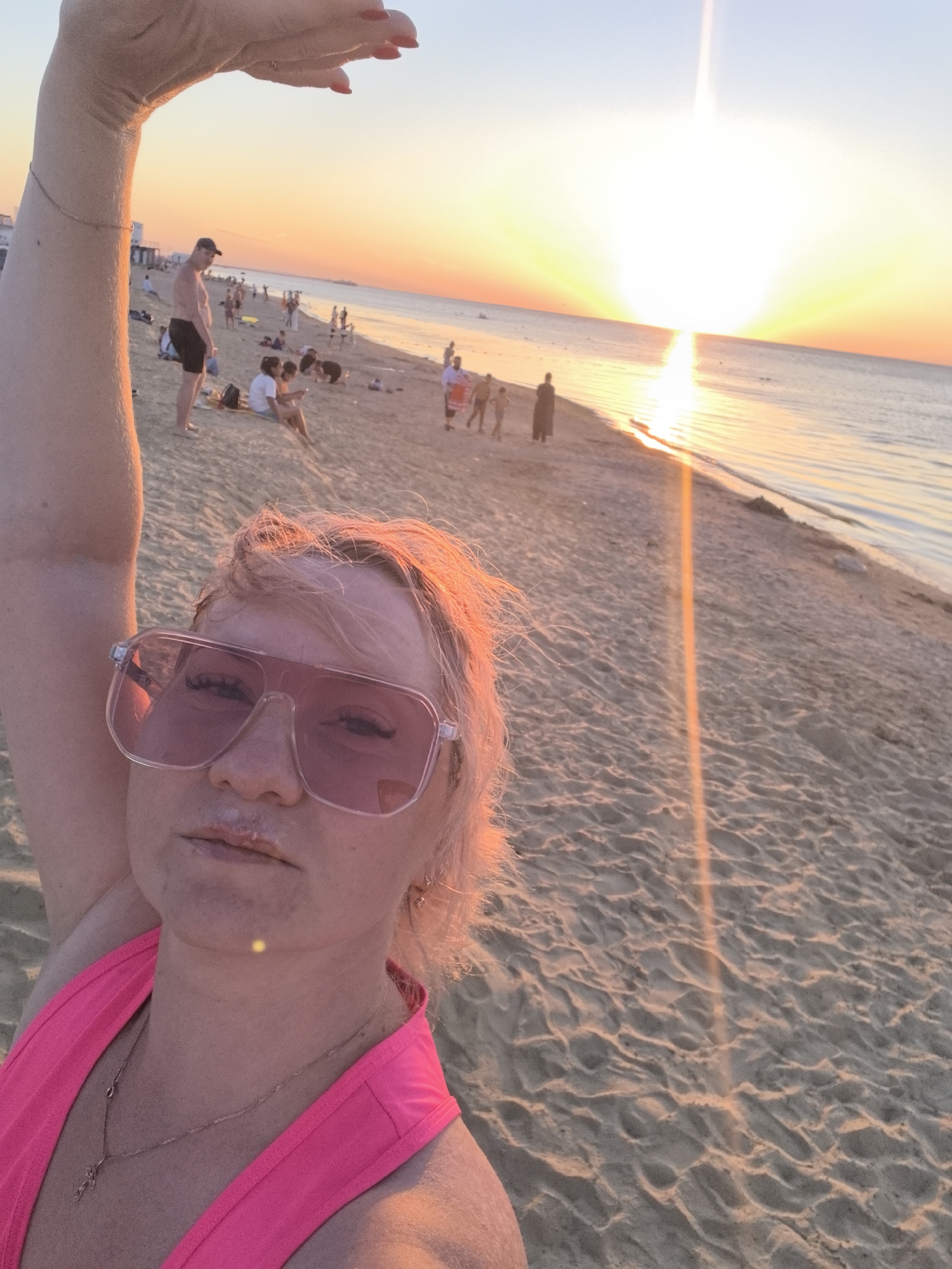 A woman takes a selfie on a beach in Anapa, Russia, at sunset. She has blond hair, is wearing large pink sunglasses and a pink swimsuit. Her left arm is raised above her head. In the background, other beachgoers are visible, as well as the Black Sea and the sun setting over the water. The sand is golden, and the sky transitions from a light blue to orange and pink hues.