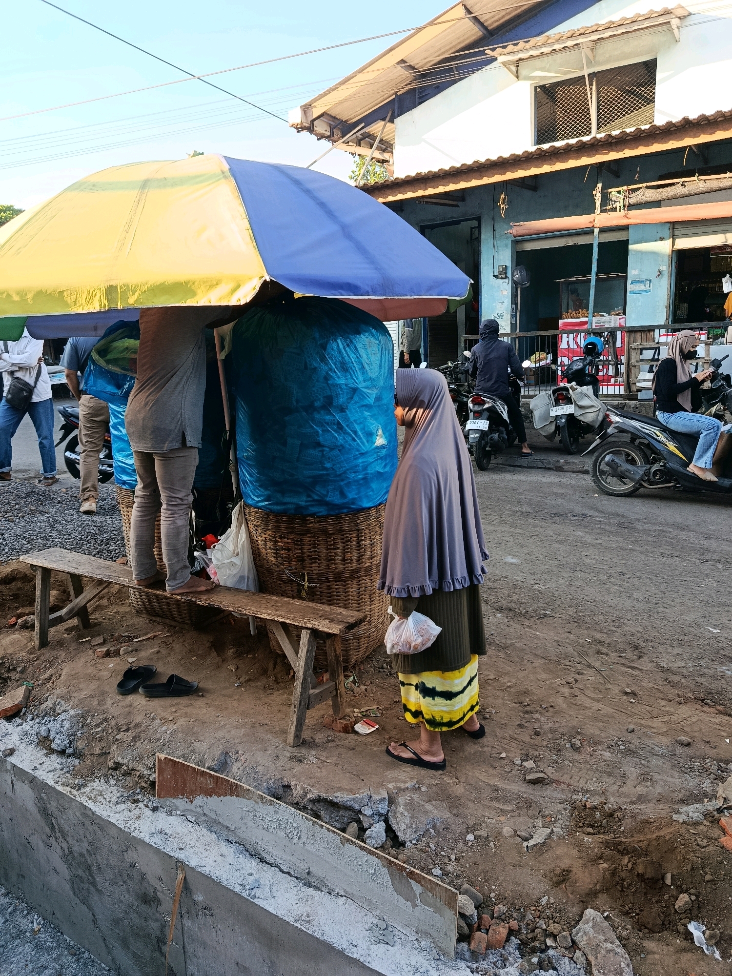 In Bululawang, Indonesia, a street scene unfolds under a bright sky, likely late afternoon or early morning due to the long shadows. A blue and yellow umbrella shades a stall where two individuals are partially visible. One person, wearing a grey long-sleeved shirt and light-colored pants, stands on a wooden bench, barefoot. Another person, mostly obscured, wears jeans and a white shirt with a dark bag slung over their shoulder. In front of the stall, a woman in a grey hijab and a yellow and black striped skirt holds a plastic bag of goods. She wears black flip-flops.
The stall itself is elaborate, featuring a large woven basket filled with what appears to be large, blue-wrapped items. A second, smaller woven basket is also present. The ground is unpaved and appears to be undergoing some form of construction, with visible trenches, piles of dirt, and cement. A rough wooden bench is positioned in front of the stall, with a pair of black flip-flops resting nearby on the ground.
In the background, a building with a light blue facade and a corrugated roof stands. Several motorcycles are parked in front of it, with a rider in a black jacket on one. Another woman, also wearing a hijab and jeans, sits on a motorcycle to the right, looking at her phone. The general environment suggests a busy roadside market or commercial area in a developing or rural part of the city.