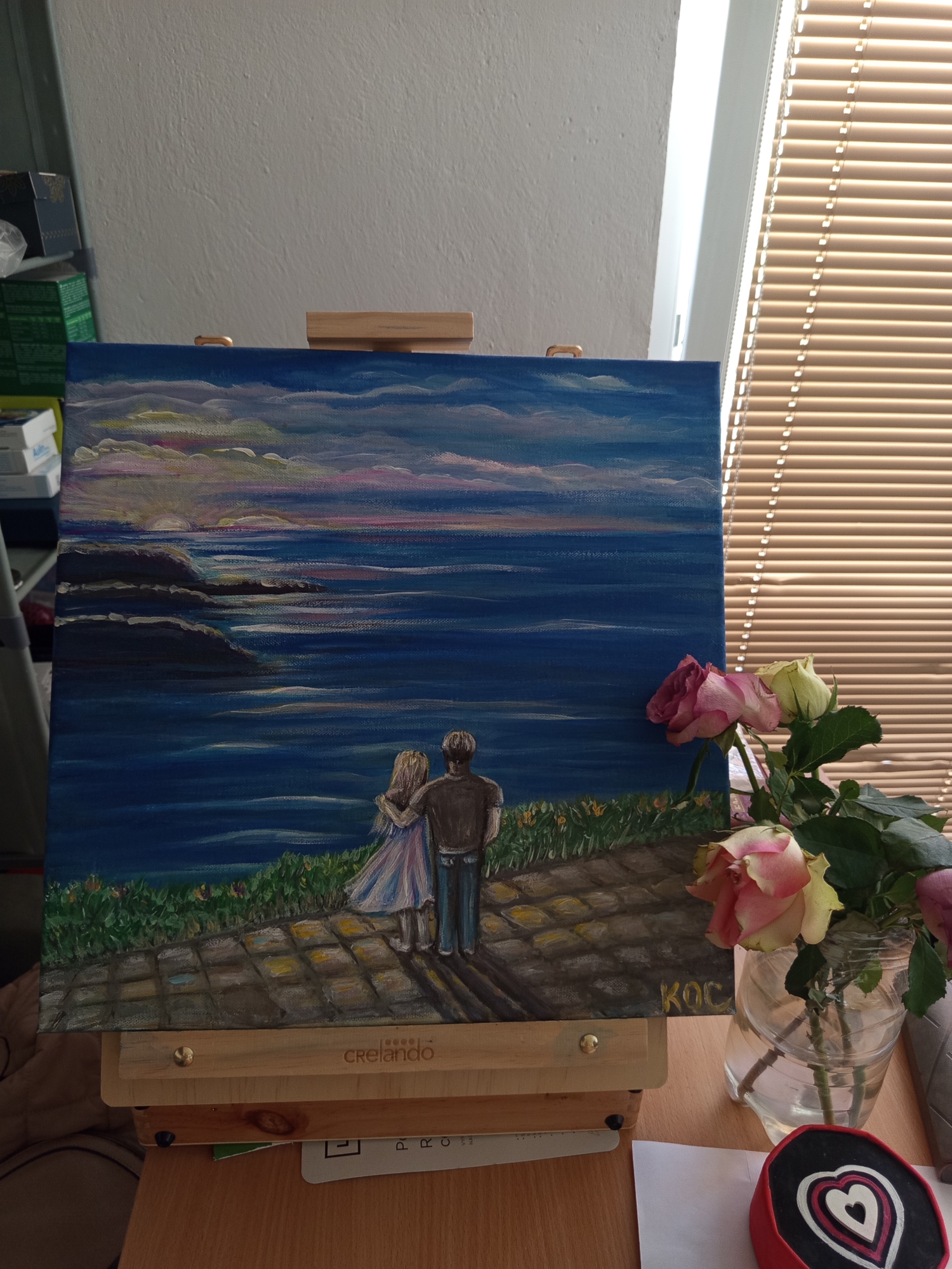 A painting of a couple embracing while looking out at a sunset over the ocean is displayed on a wooden easel. The painting depicts a rocky coastline with the sea stretching out to the horizon. The sky is filled with colorful clouds, and the sun is setting, casting a warm glow over the scene. The couple, facing away from the viewer, are positioned on a paved path overlooking the water. Green grass and small yellow flowers grow along the edge of the path. To the right of the painting, a bouquet of pink and yellow roses in a glass jar sits on a wooden table. A small red object with concentric heart designs is also visible on the table. The background shows a white wall and blinds on a window. The painting is signed with the initials "KOC". The easel has a "Crelano" logo. The scene appears to be an indoor setting, possibly a room or studio, with natural light coming from the window. There is no indication of the city of Brno, Czechia.